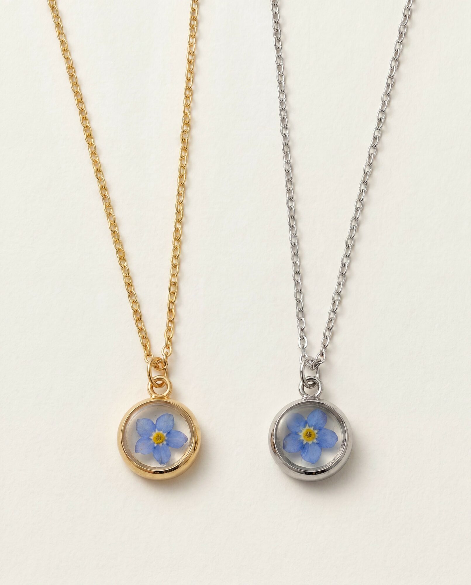 Two necklaces with round pendants containing blue flowers, one gold and one silver, on a white background.
