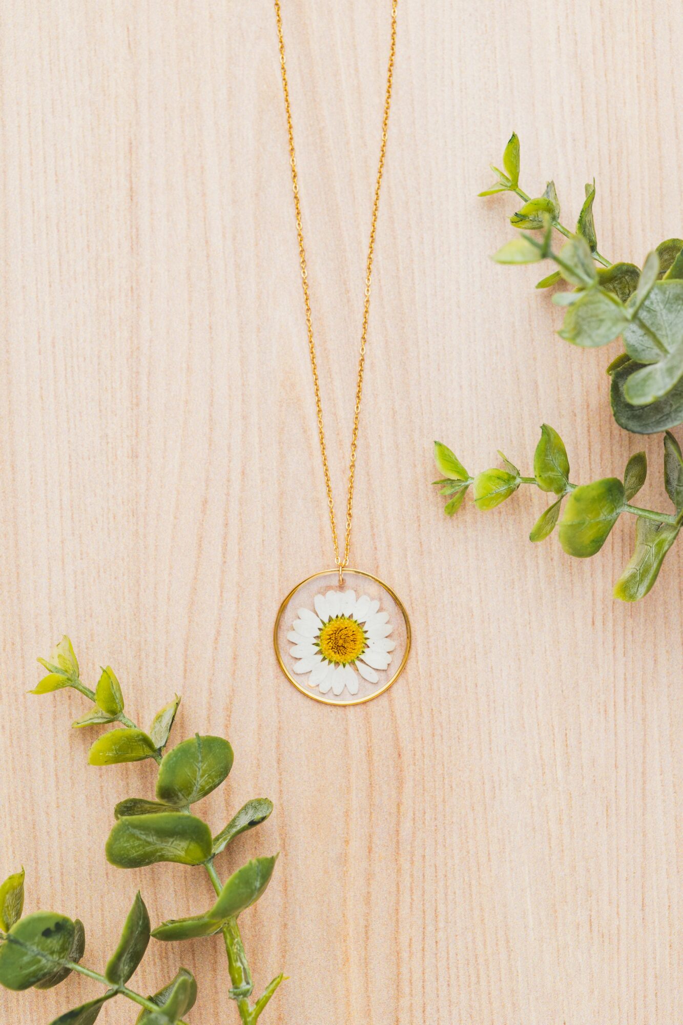 Gold chain necklace with a round pendant featuring a daisy flower, placed on a light wooden surface with green leaves.