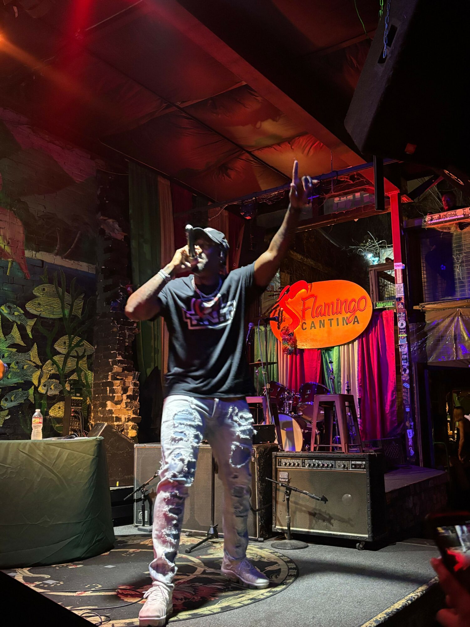 Person on stage holding microphone, performing with colorful background and neon sign, stage equipment visible.