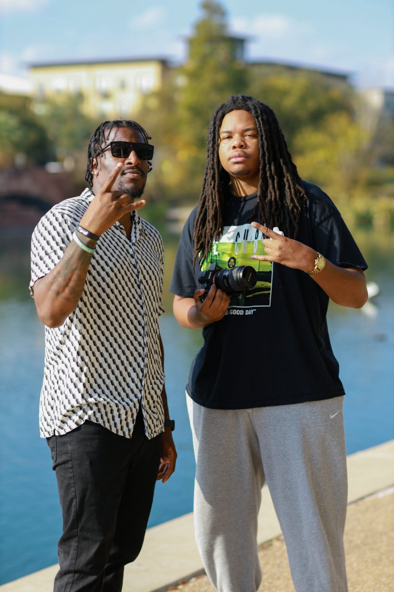 Two young men stand outdoors near water, one making a peace sign, the other holding a camera, with trees and buildings in background.