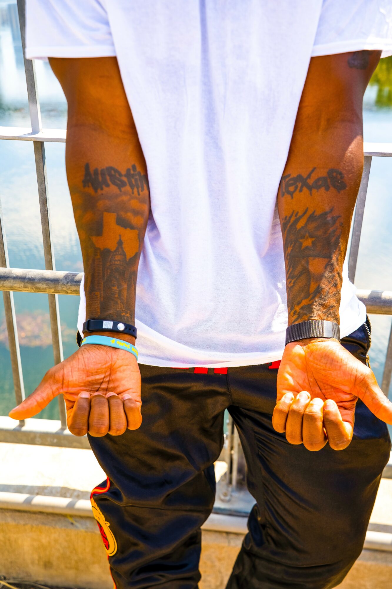 Person with tattoos on arms, wearing a white shirt and black pants, standing outdoors near a railing.