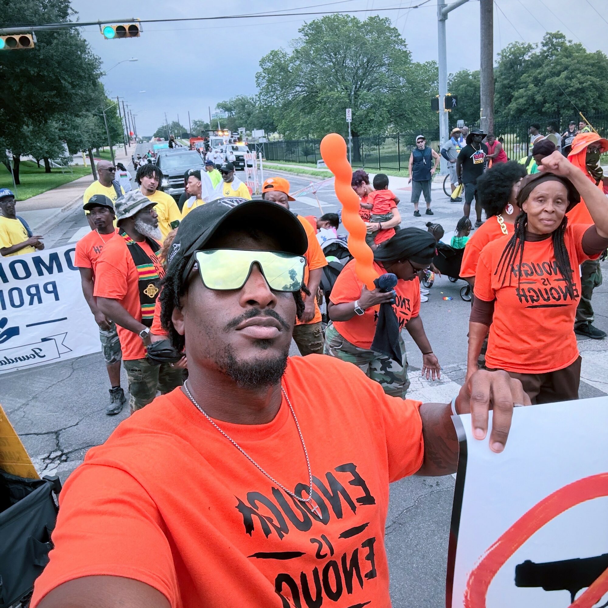 Man taking selfie at protest, surrounded by people holding signs and balloons, wearing orange shirt and sunglasses.