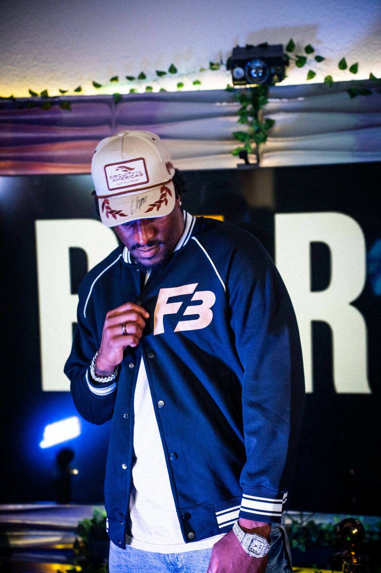 Person wearing a baseball cap and jacket, standing in front of a large sign with letters, at an event or party.