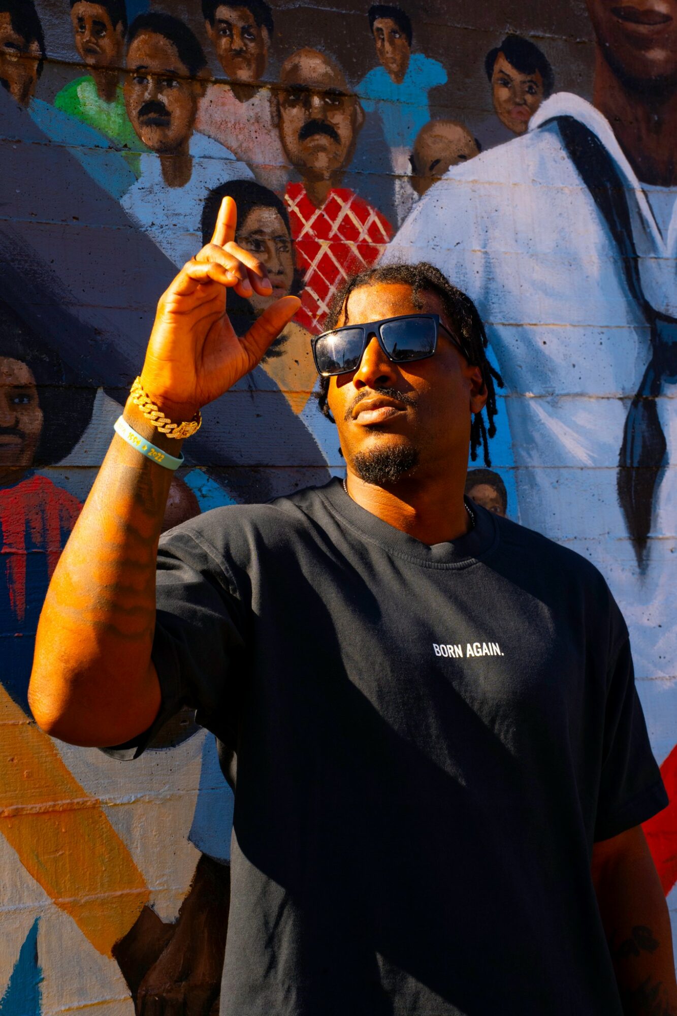 Man with sunglasses and black T-shirt making a gesture with his hand, crowd mural background.
