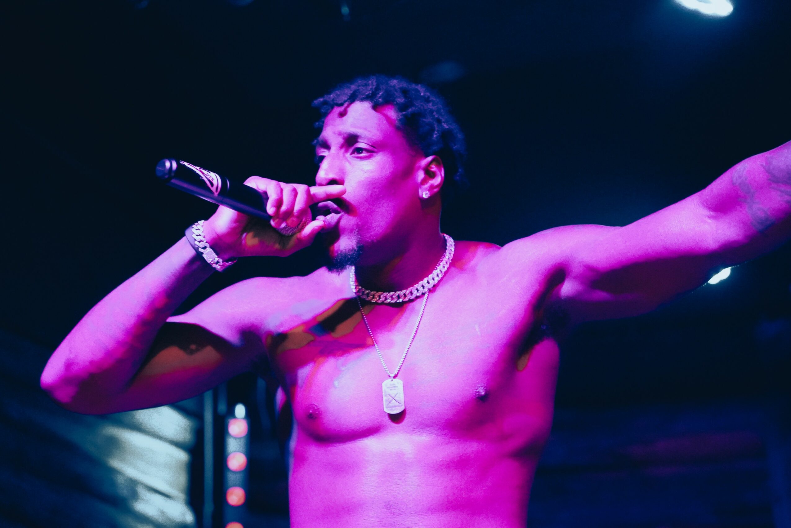 A shirtless male performer with short curly hair singing into a microphone on stage, with one arm extended.
