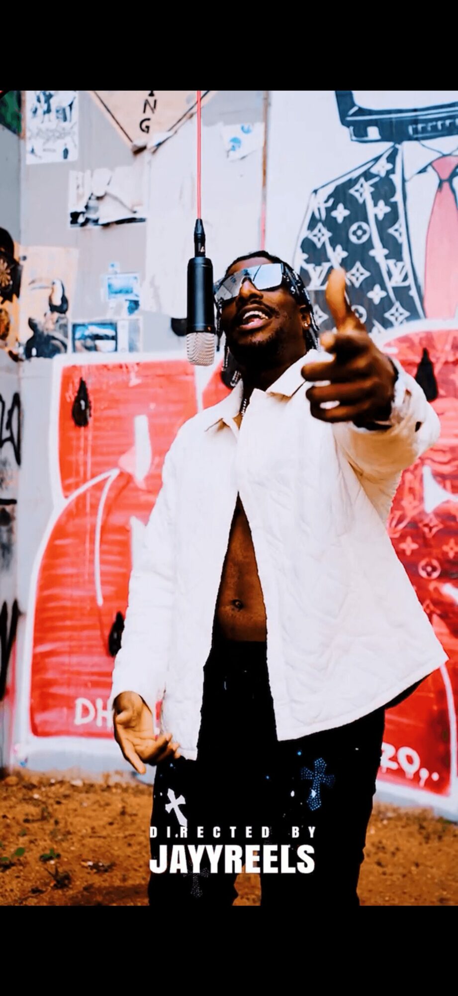 Person singing into a microphone, smiling, wearing glasses and a white jacket, in front of colorful graffiti, giving a thumbs-up.