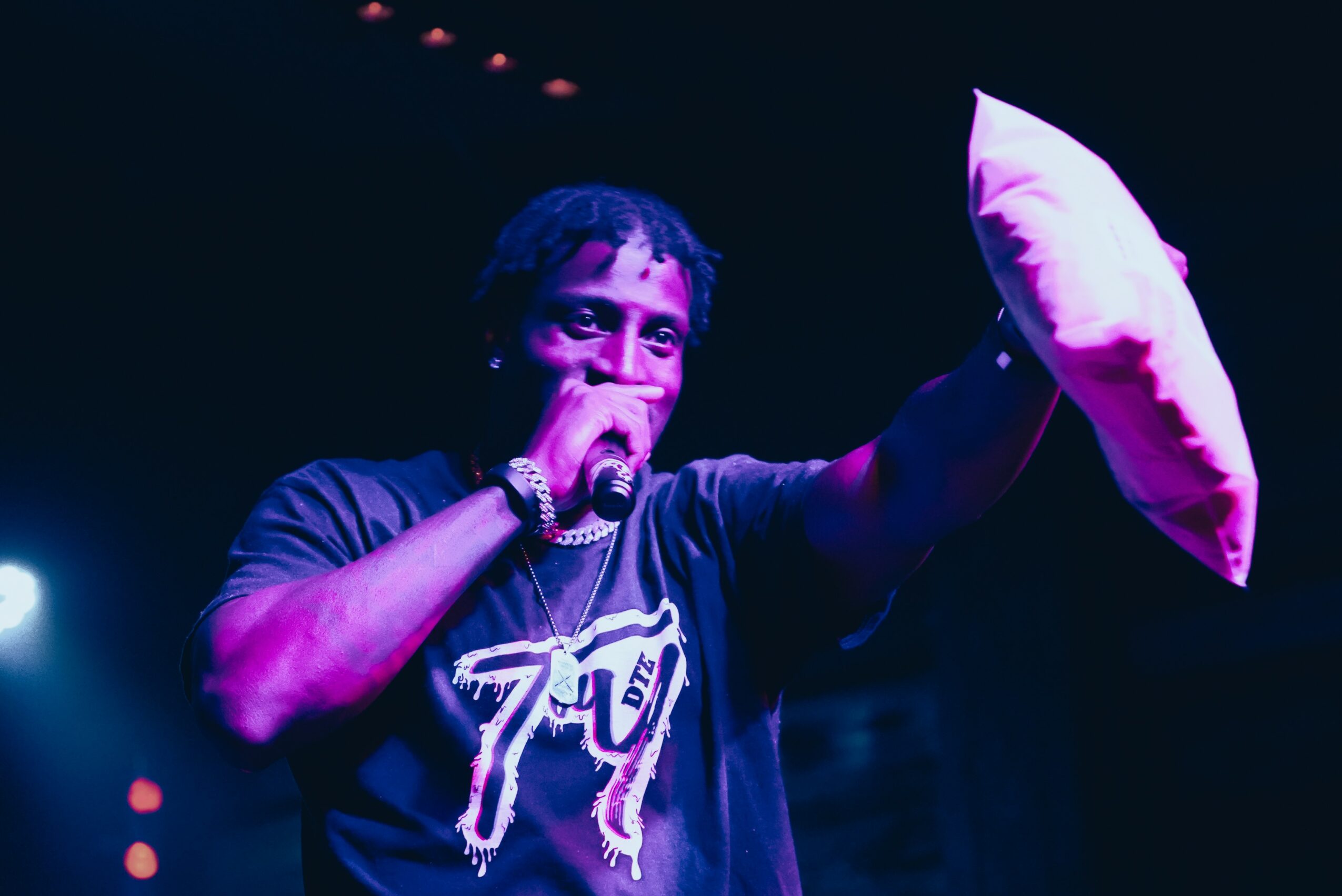 Person holding a pillow in one hand and a microphone in the other, on stage with dark background and stage lights.