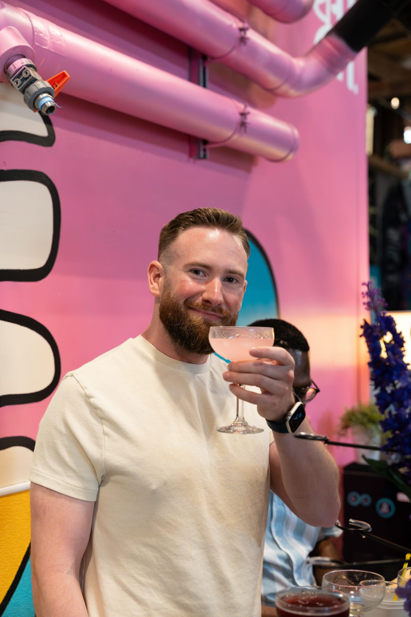 Man with beard holding a glass, smiling, in front of pink pipes and a pink background, celebrating.