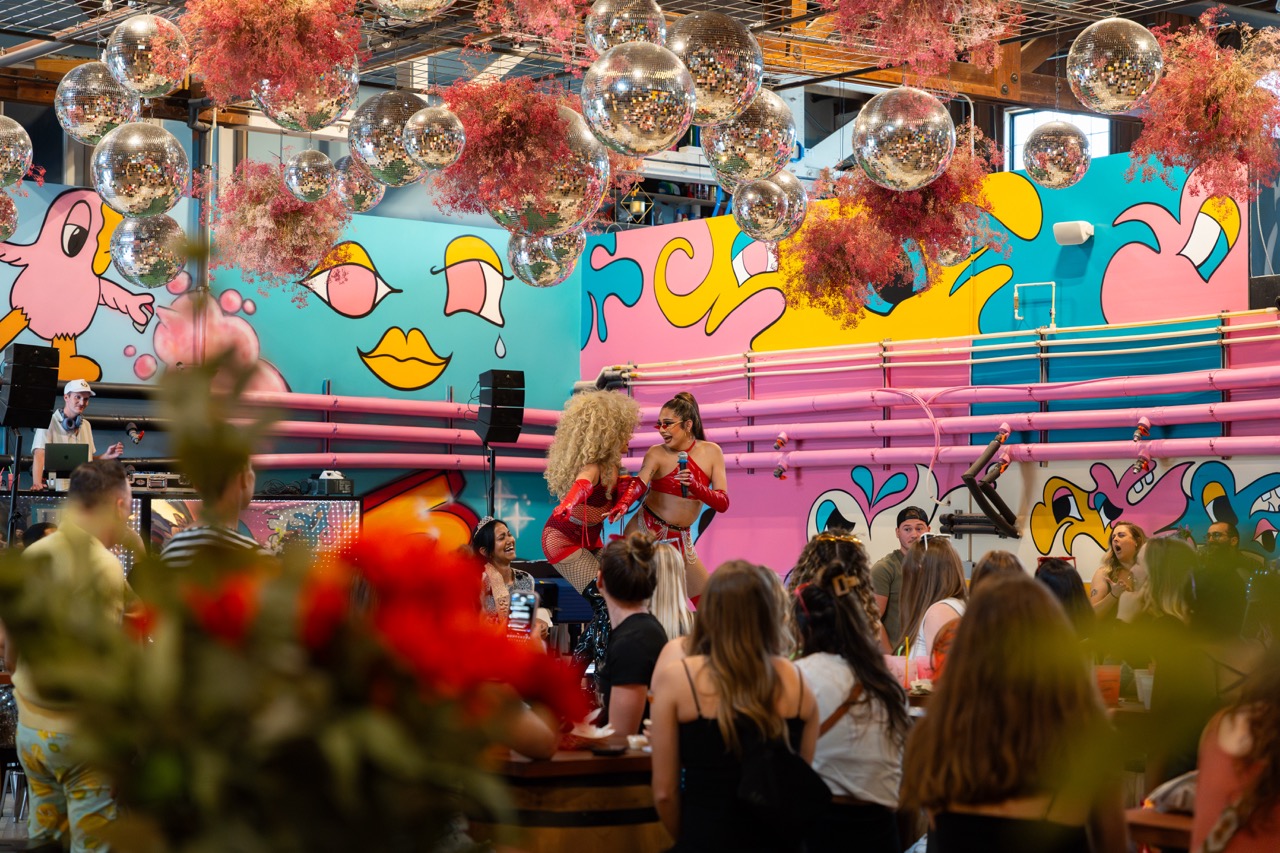 People gather in front of a colorful mural with abstract faces, pink and blue patterns, and hanging decorations, including balloons and flowers.