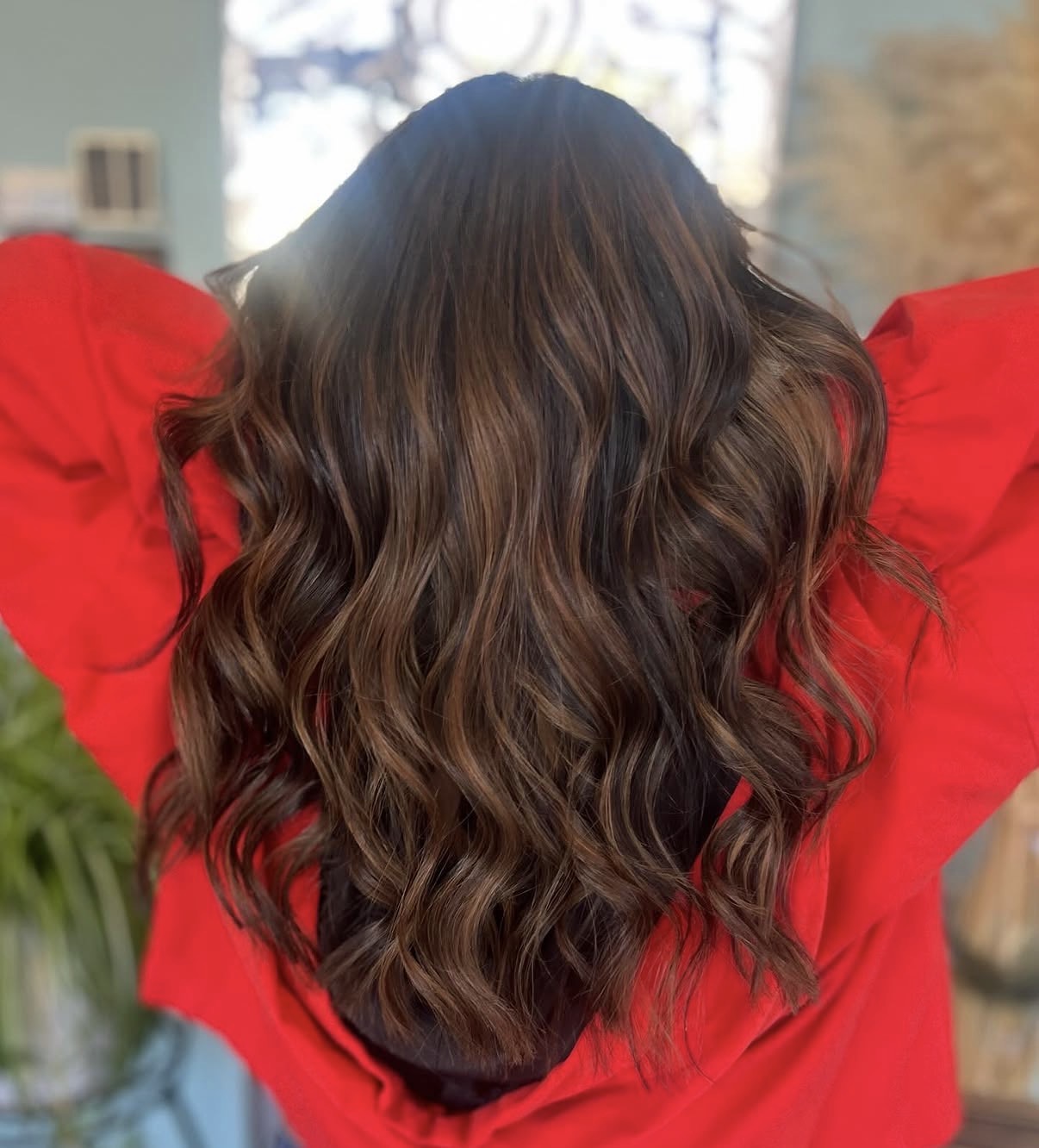 Back view of a person with long, wavy brown hair wearing a red top, outdoors with trees and building in background.