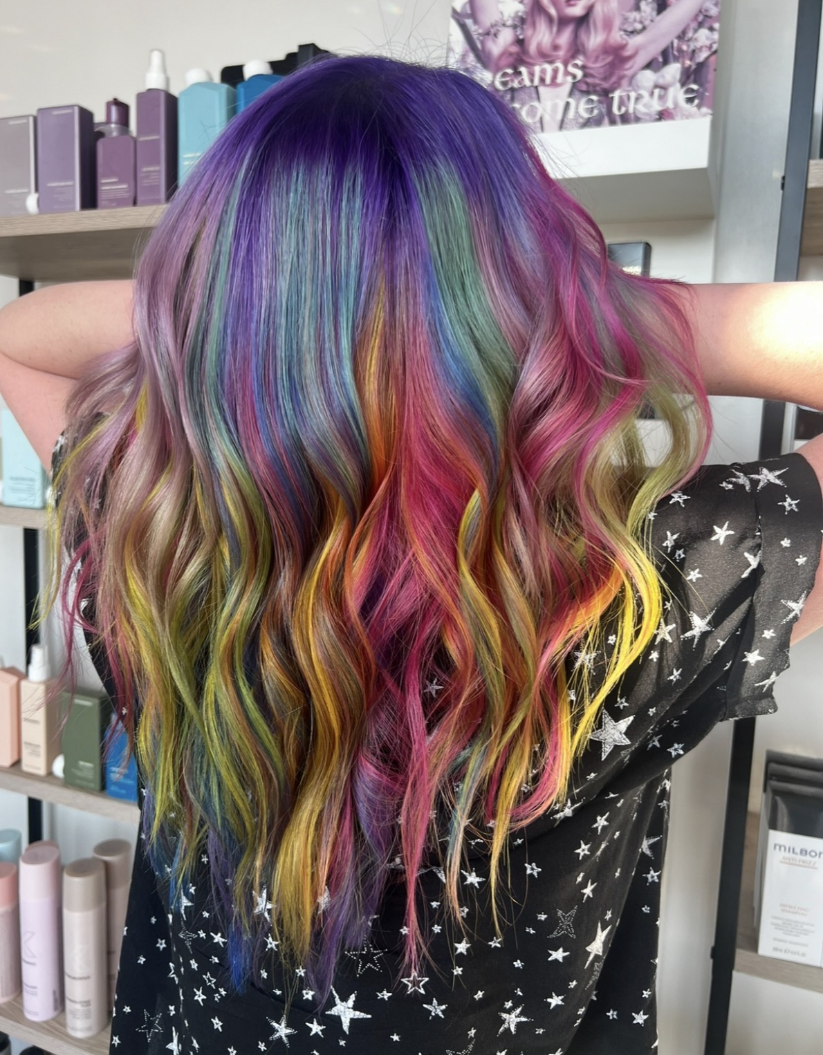 Person with long, wavy, rainbow-colored hair, standing in front of shelves with hair products, wearing a star-patterned shirt.