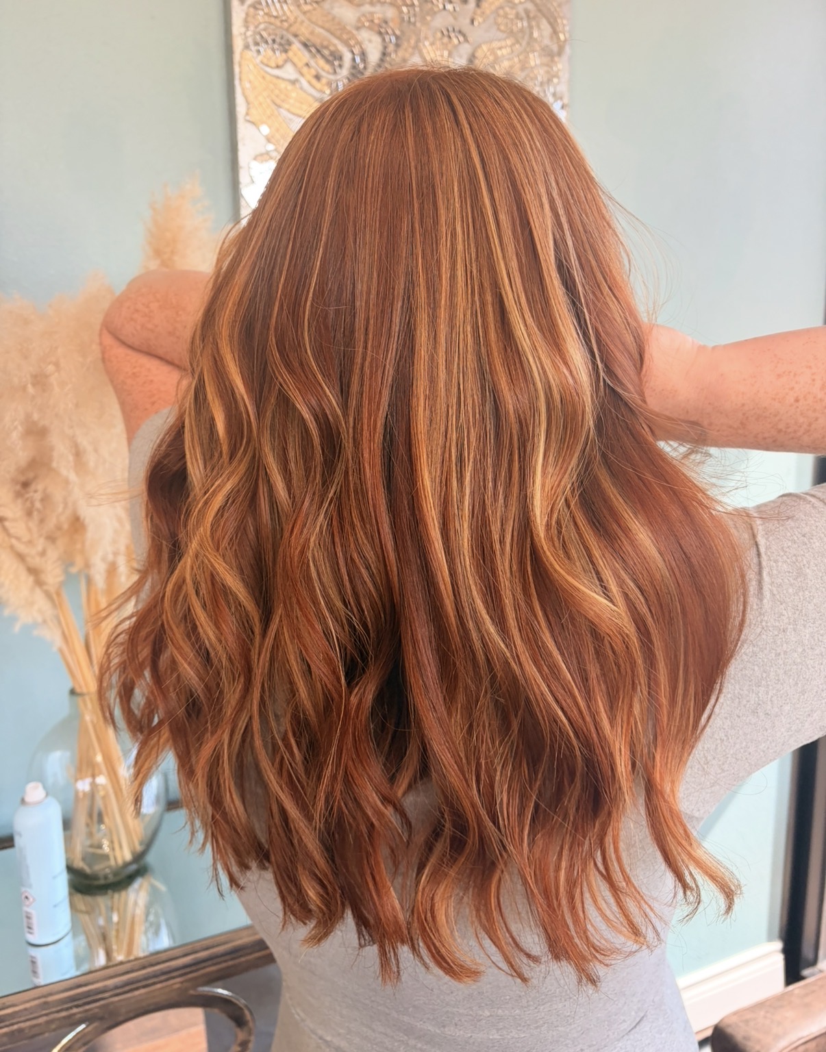 Back view of a person with long, wavy, reddish-brown hair in a room with light-colored walls and decor.