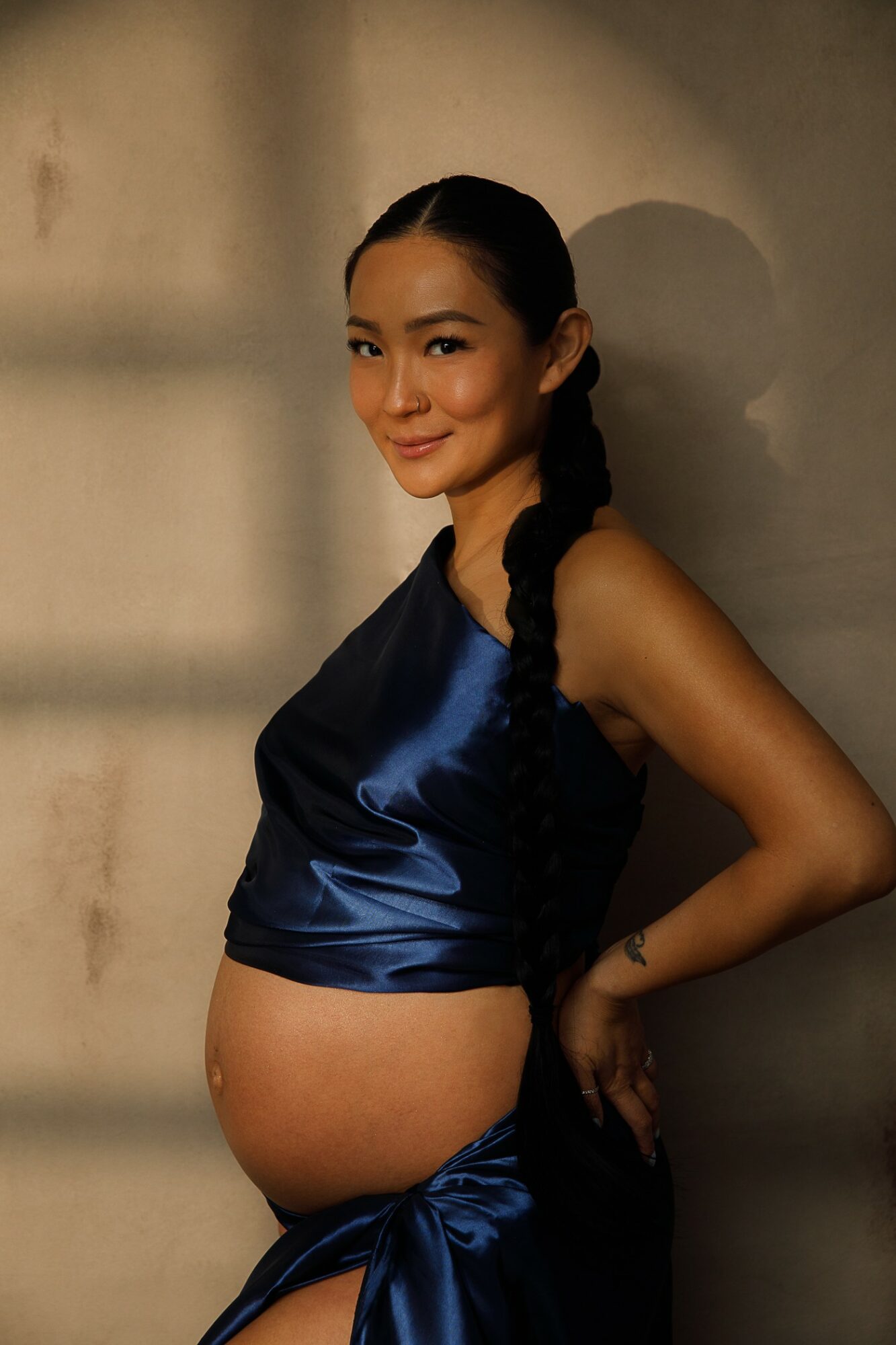 Pregnant woman with long braided hair wearing a dark blue satin top and matching skirt, standing against a wall.