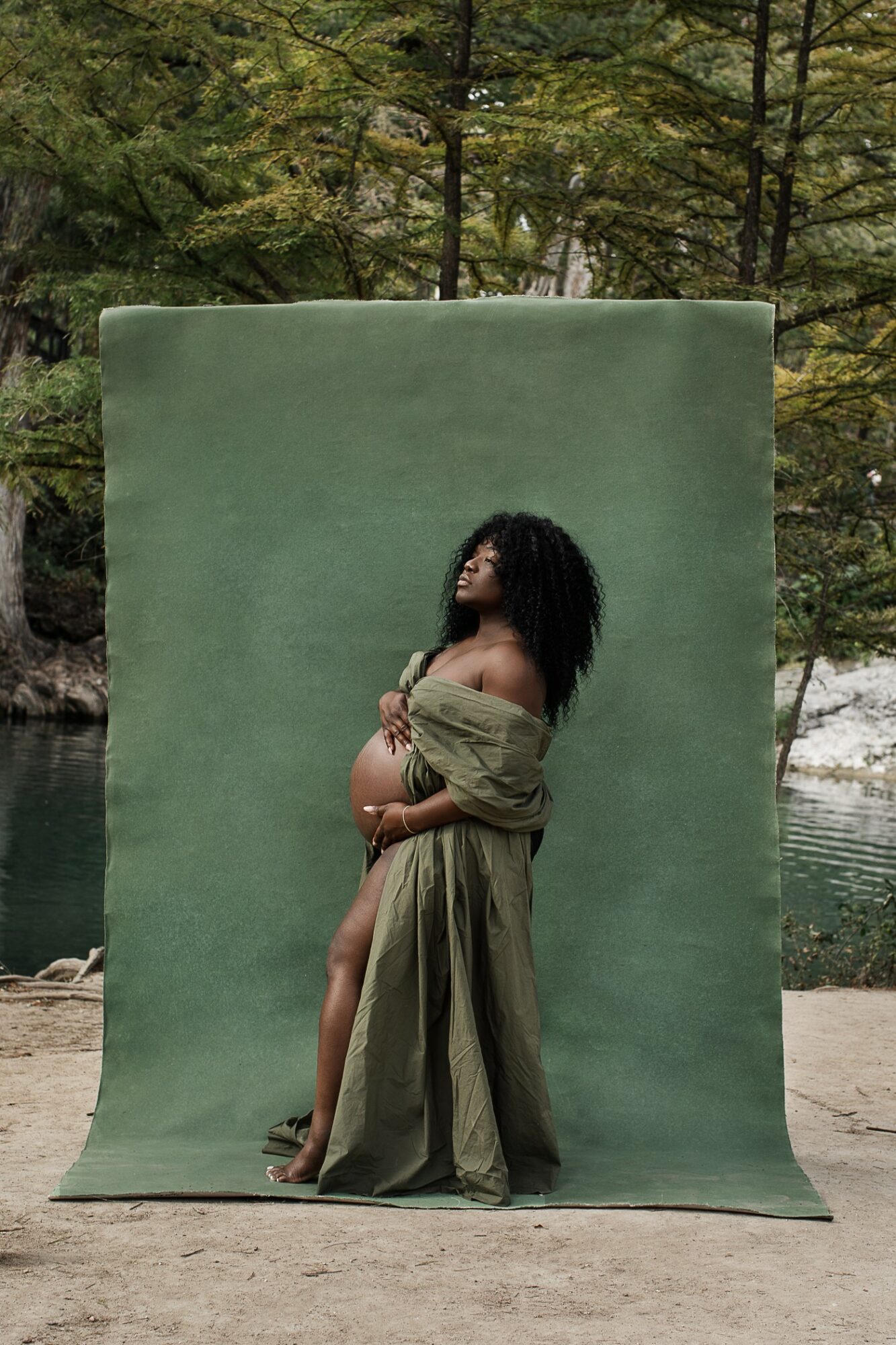 Pregnant woman standing outdoors in front of a green backdrop, wearing a long dress, with trees and water in the background.