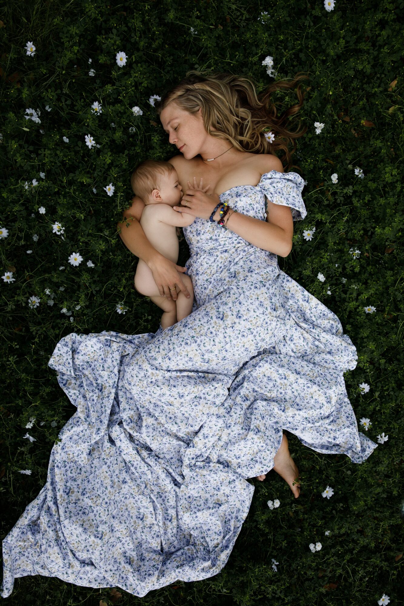 Two women lie on grass with small white flowers, one holding the other, both smiling and looking at each other.