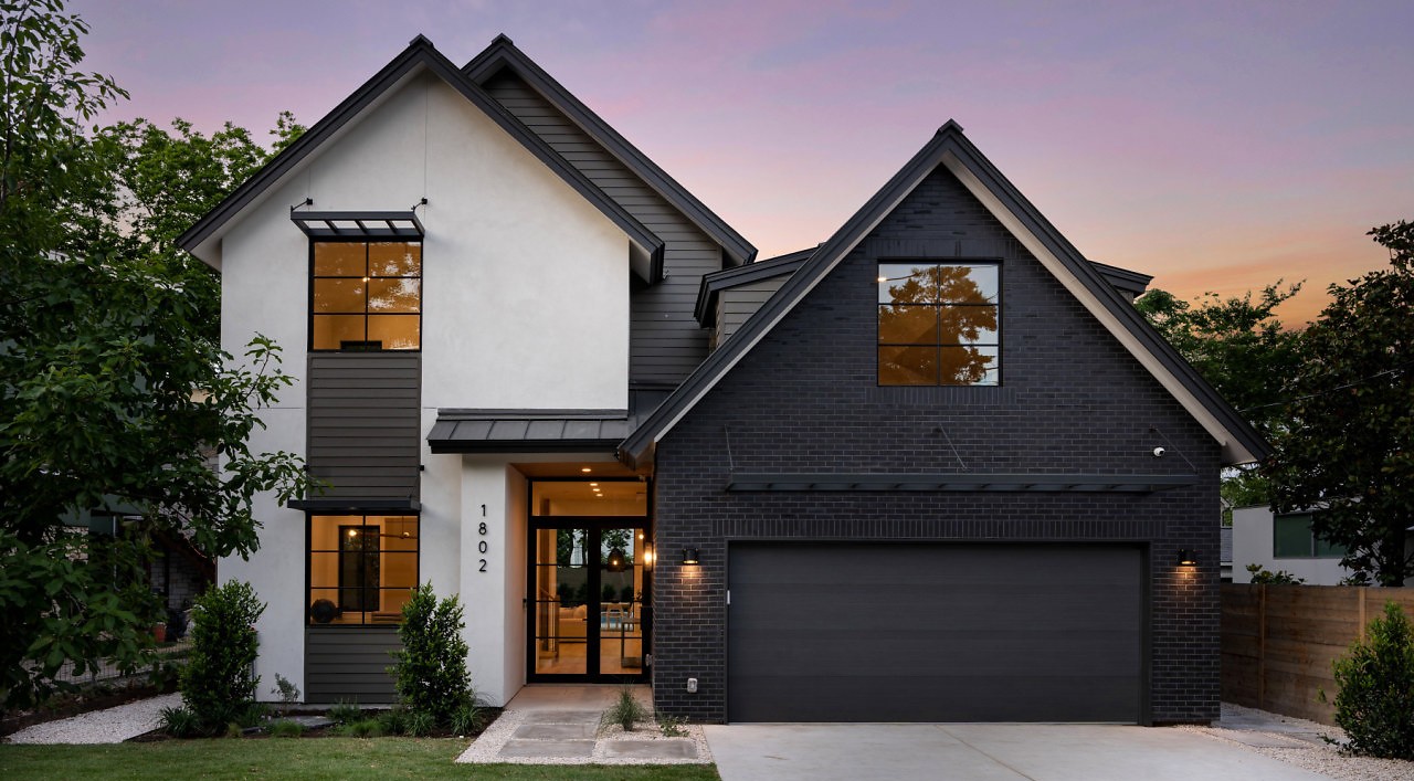 Modern two-story house with white and black exterior, large windows, and a two-car garage, surrounded by trees and a lawn.