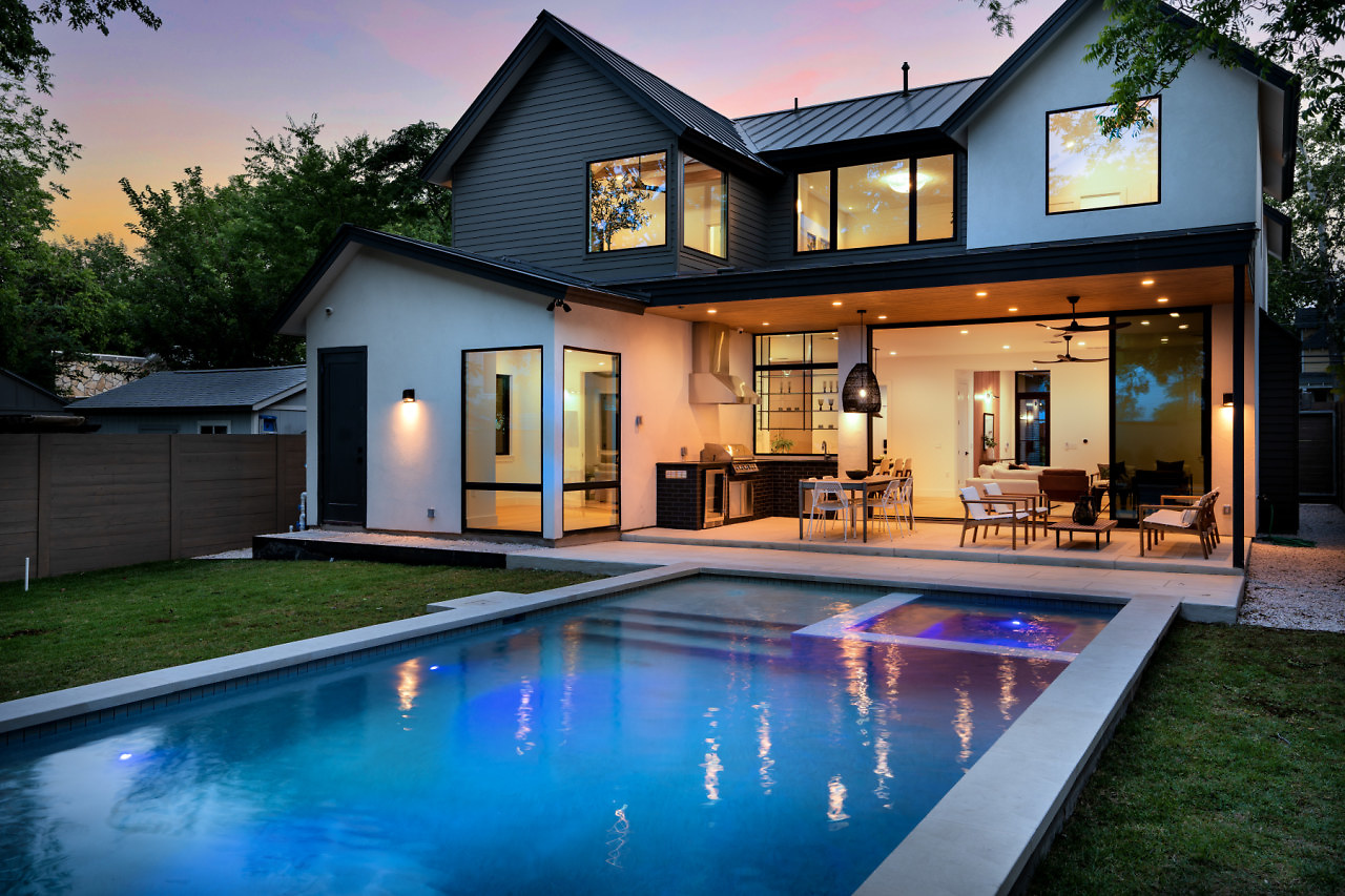 Modern two-story house with large windows, outdoor patio, and swimming pool in backyard during evening, with trees and sky in background.