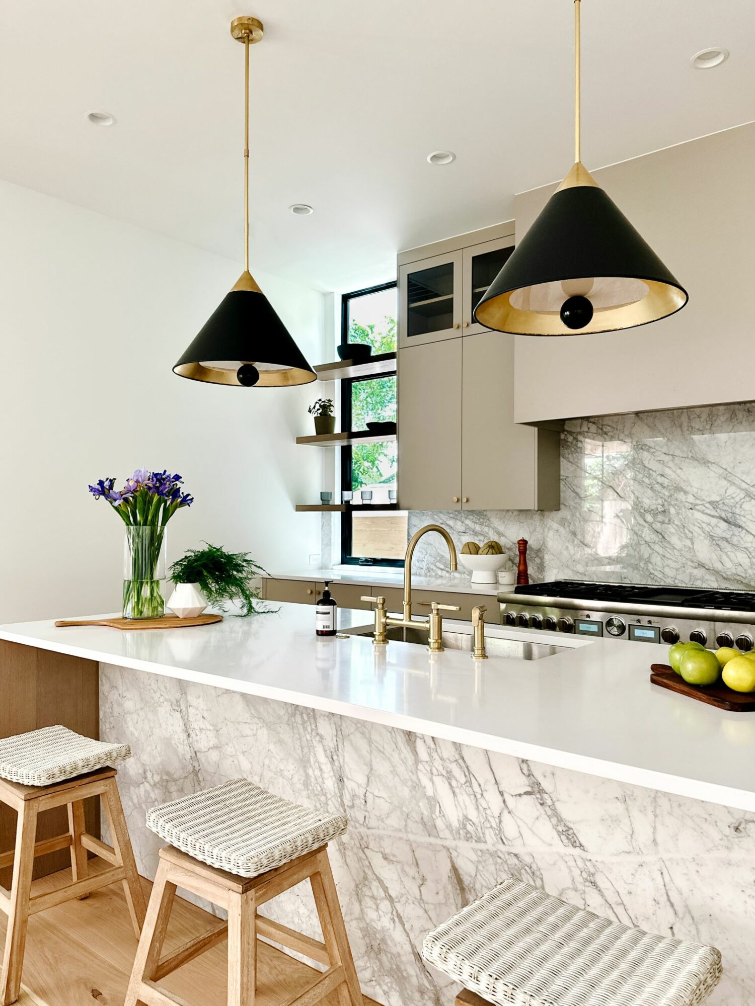 Modern kitchen with white marble island, black pendant lights, and a window with open shelves, flowers, and apples.