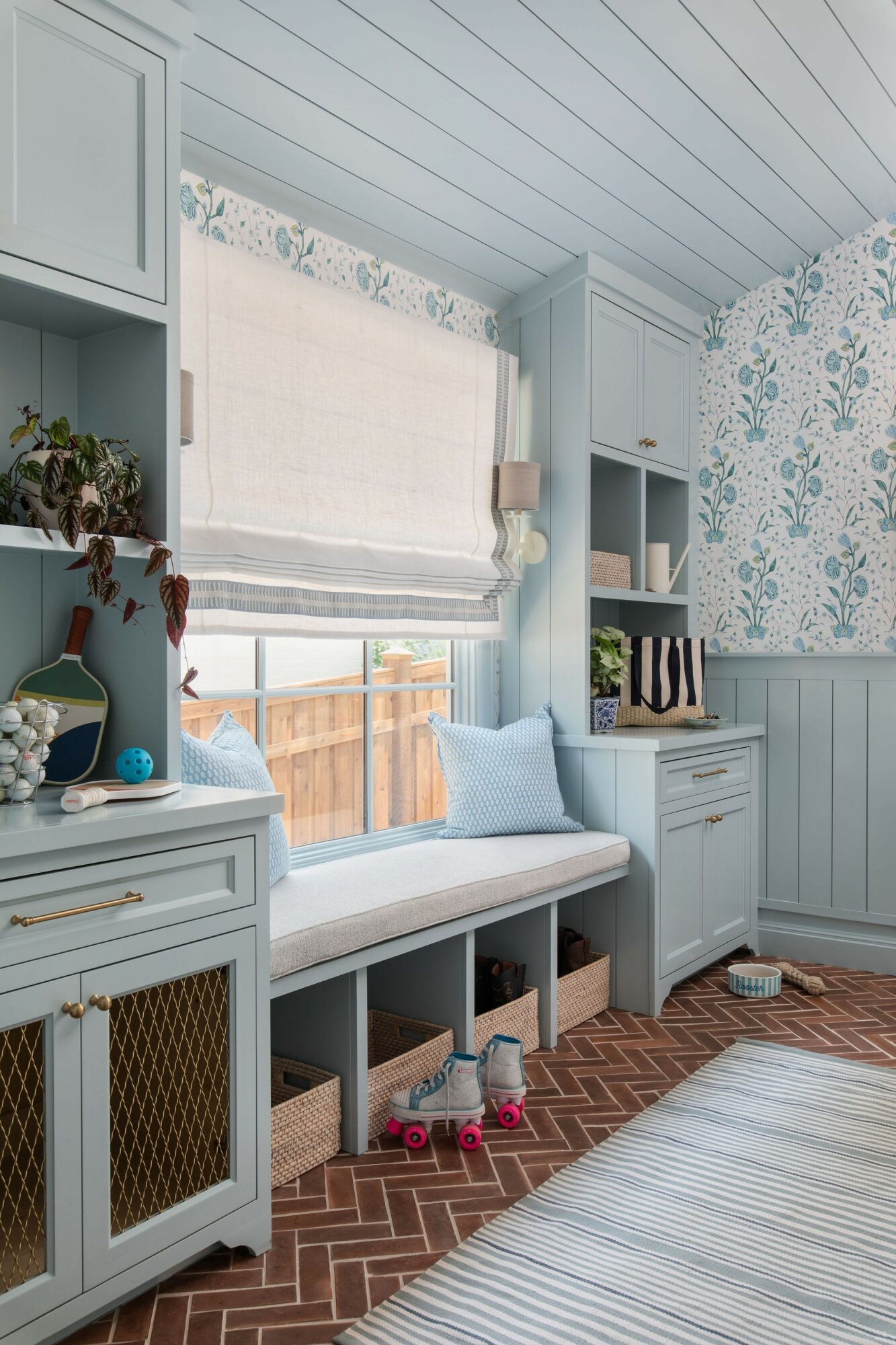 Cozy window seat with cushions and storage, light blue cabinetry, floral wallpaper, and a window with a Roman shade.