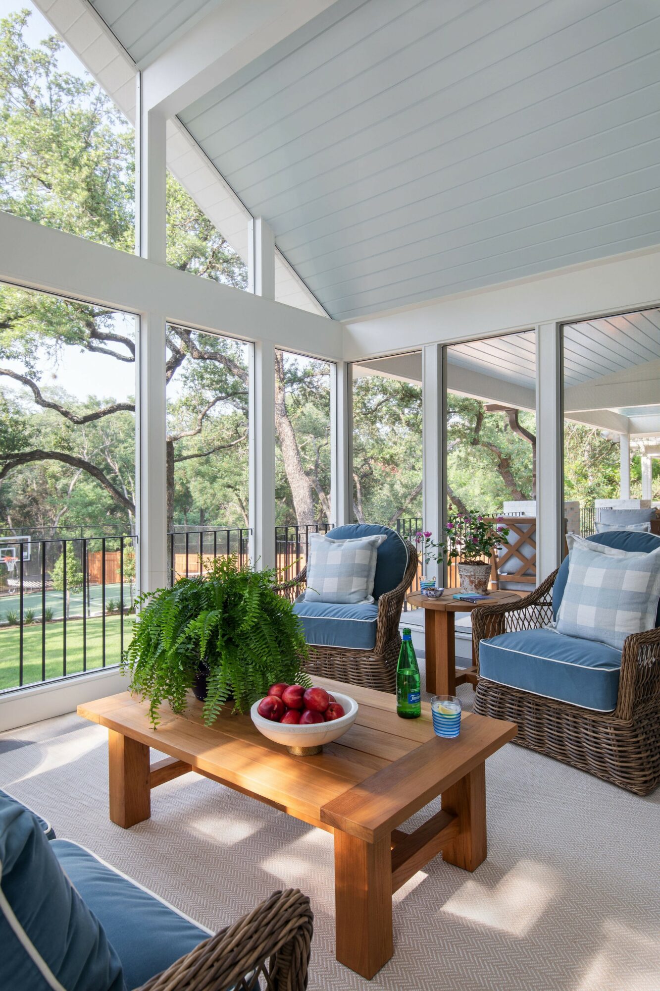 Sunlit porch with wicker chairs, a wooden table, and large windows showing trees outside.