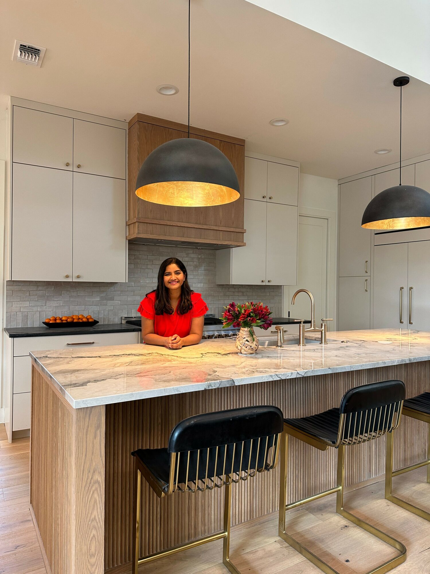 Woman in red top sitting at kitchen island with black chairs, pendant lights, and white cabinets in background.