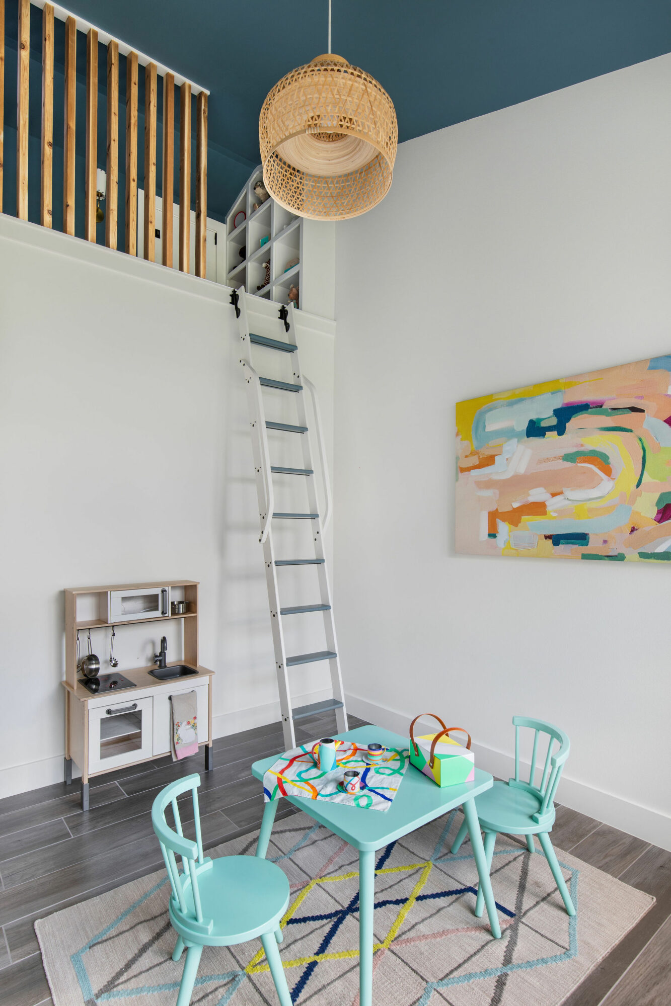 Children's playroom with a small table, four chairs, a ladder, and colorful wall art, with a hanging wicker basket and a railing above.