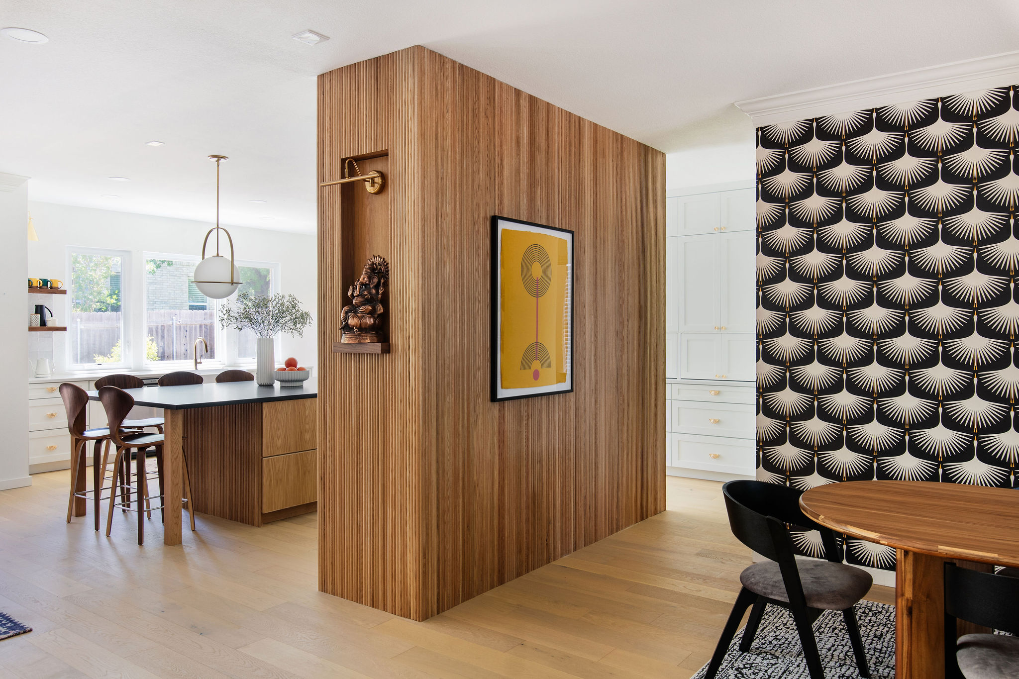 Wood-paneled wall with framed artwork, patterned wall, dining table with chairs, and kitchen area in background.