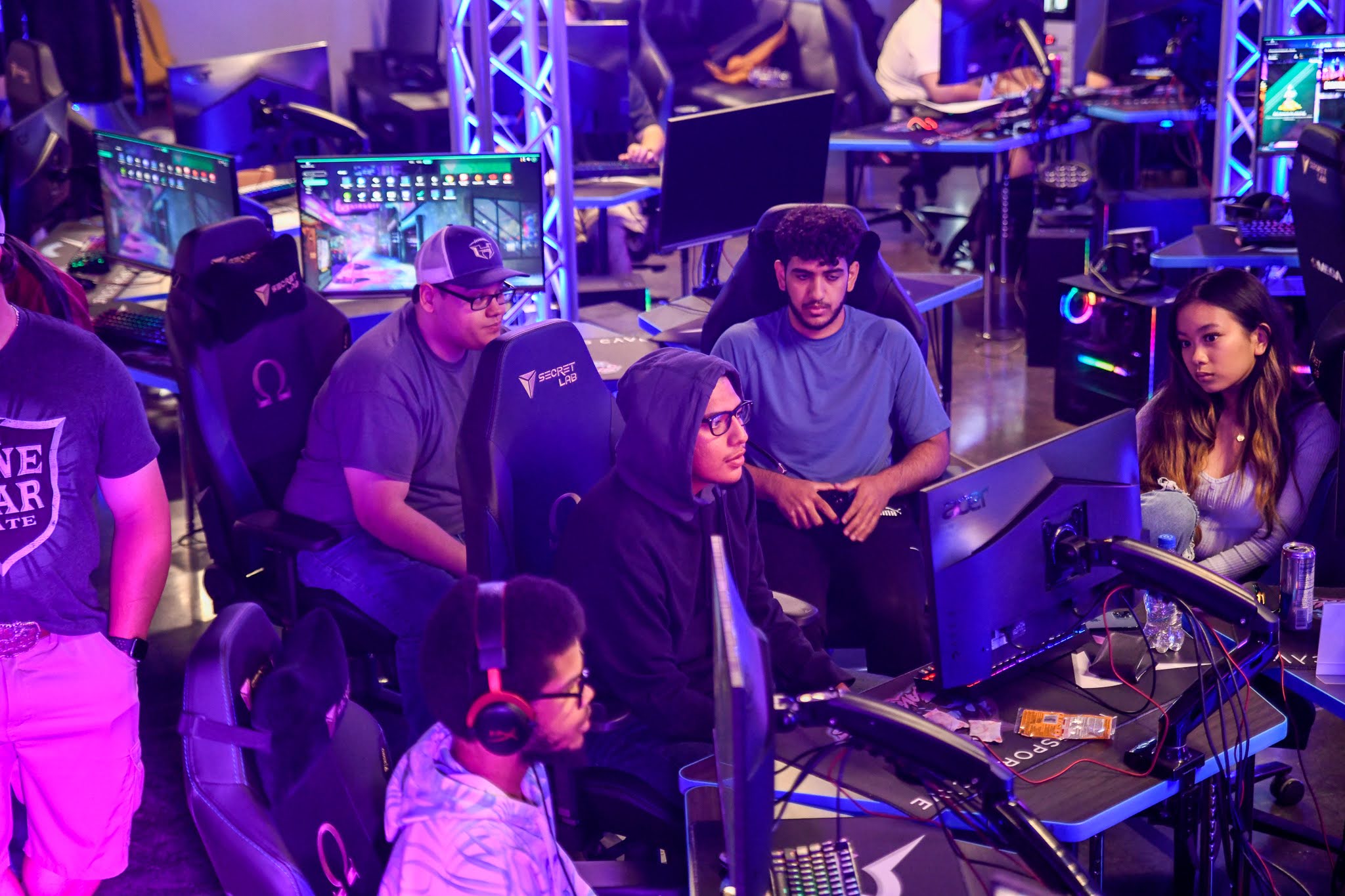 Group of people gaming at computers in a brightly lit room with screens and gaming equipment, some standing and some seated.