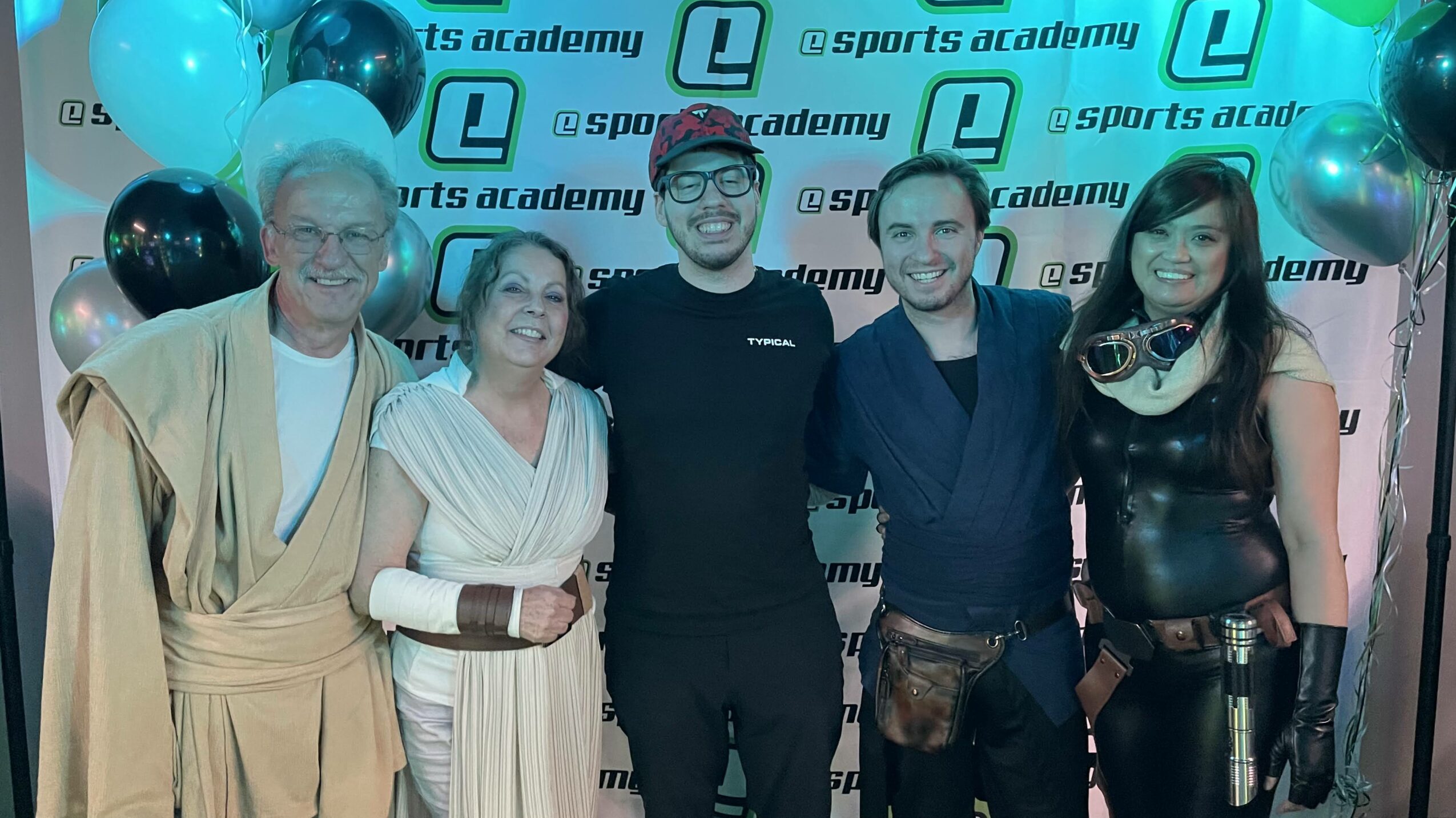 Group of five people smiling at an event with balloons and a branded backdrop.