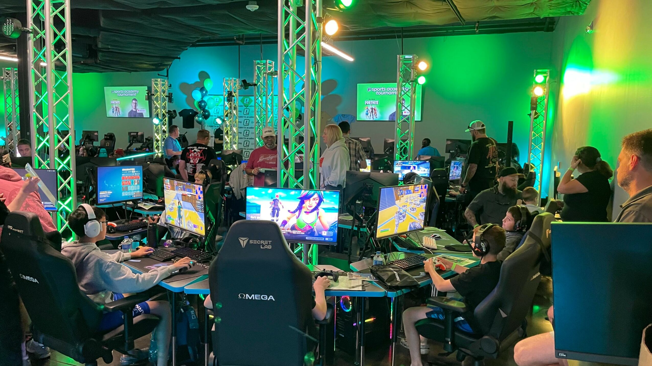 Gaming event with people playing on computers, colorful lighting, and screens displaying games, in a large room with green and yellow lights.