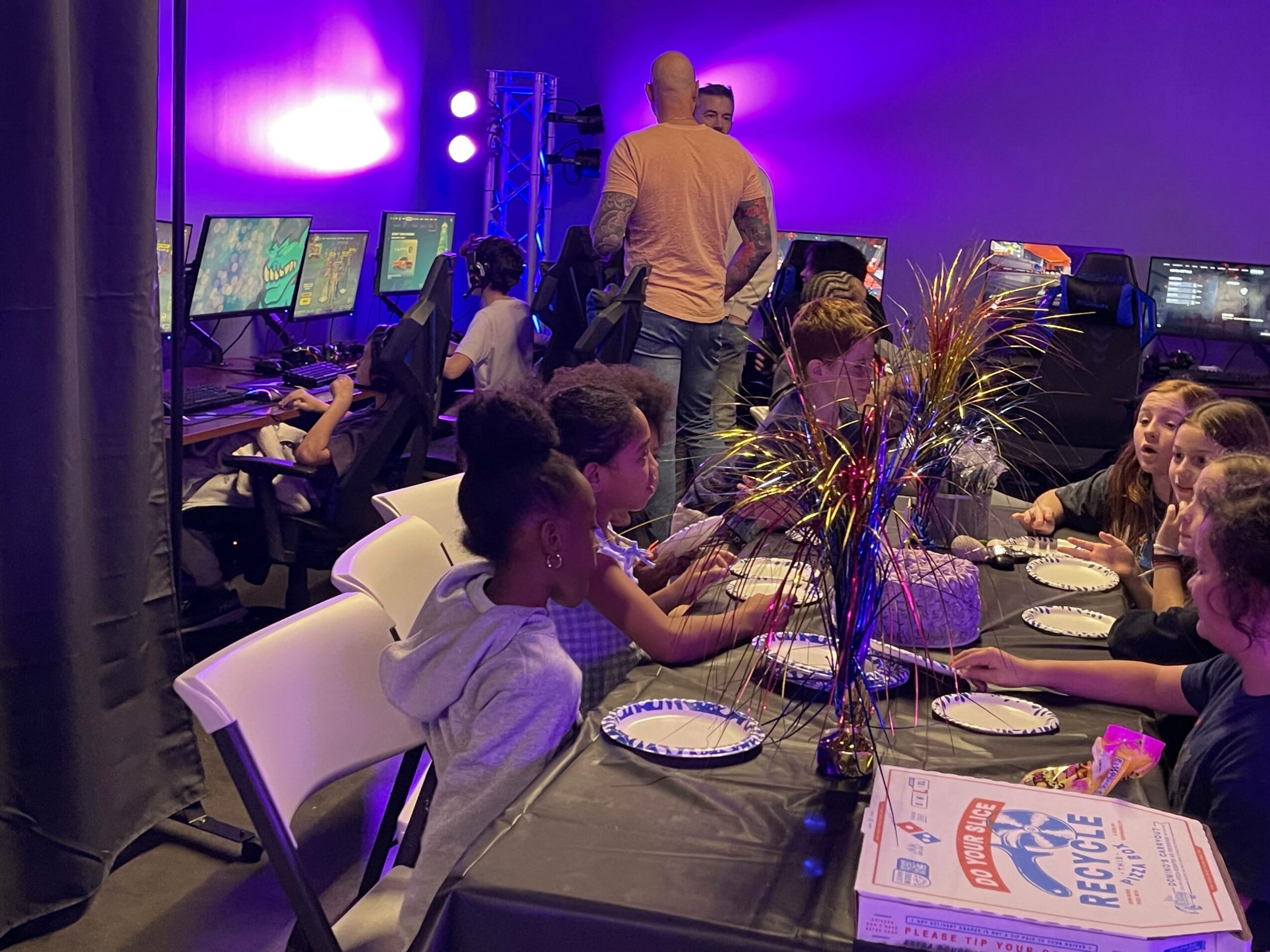 People seated at a table with plates and a large flower arrangement, gaming monitors and people in the background, purple lighting.