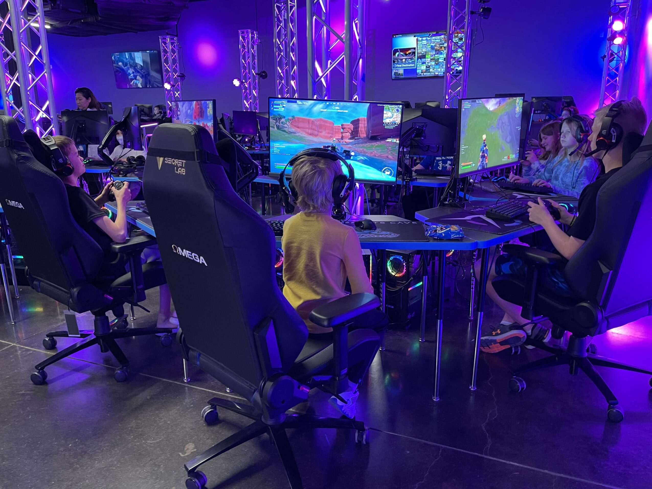 Children playing video games on screens in a room with purple and blue lighting, seated in gaming chairs.
