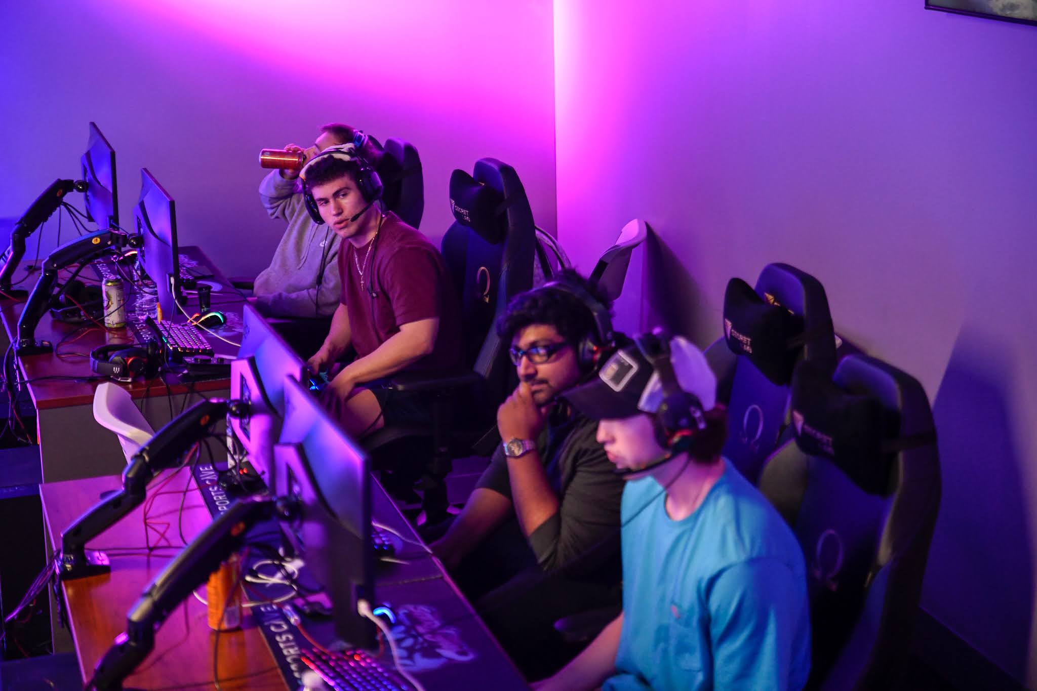 Three people sit at a gaming station with monitors, wearing headphones, in a room with purple lighting.