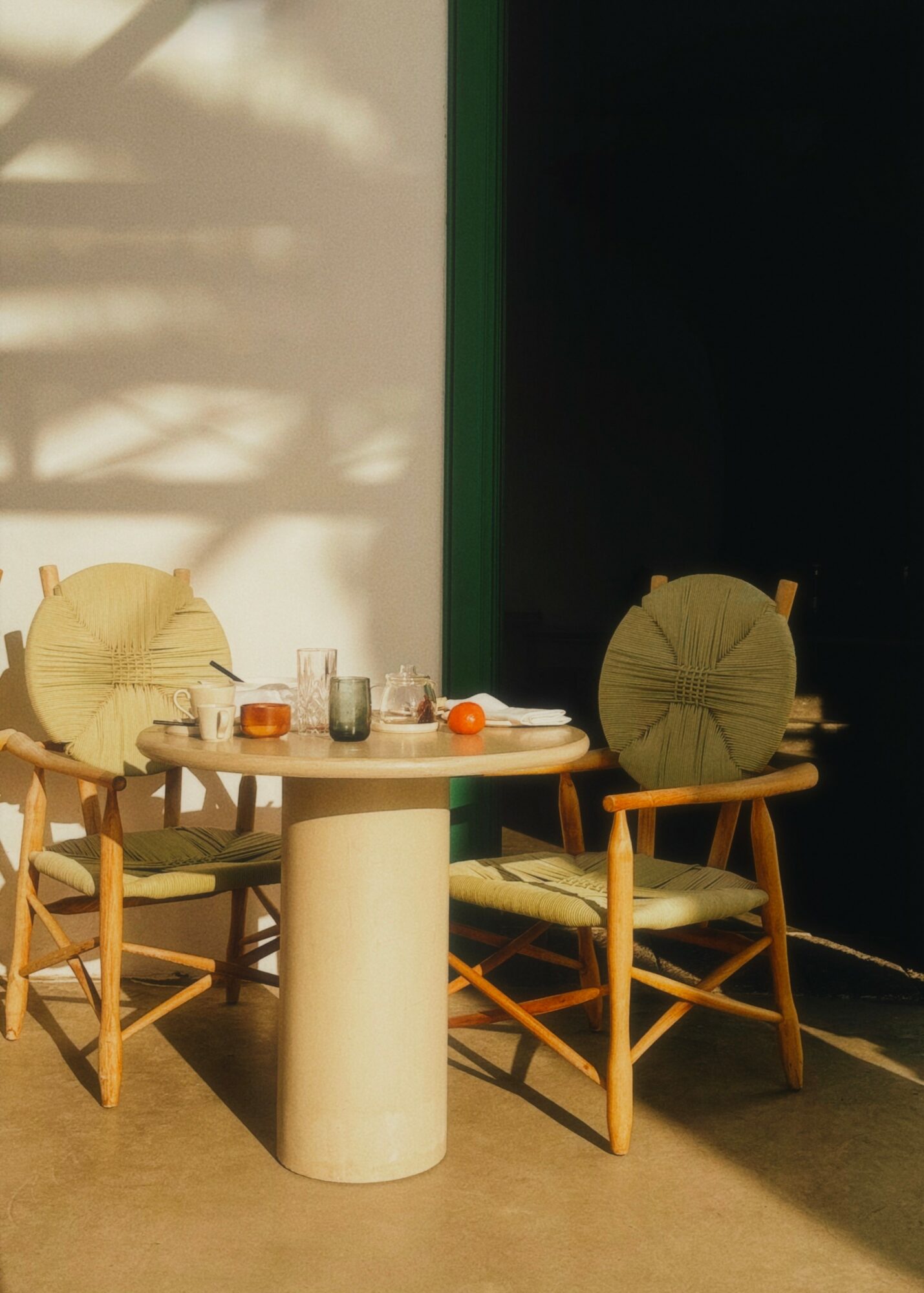 Table with four chairs, some objects on the table, sunlight and shadows, and a dark background.