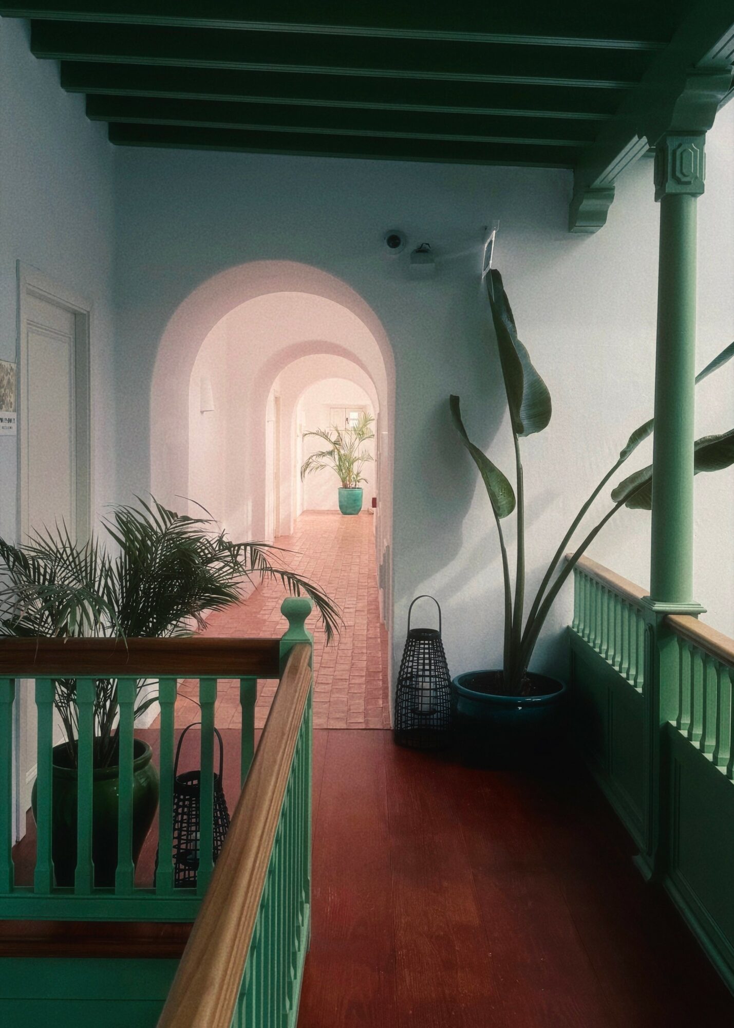 Indoor hallway with arched doorway, plants, wooden floor, and green railing, viewed from a staircase.