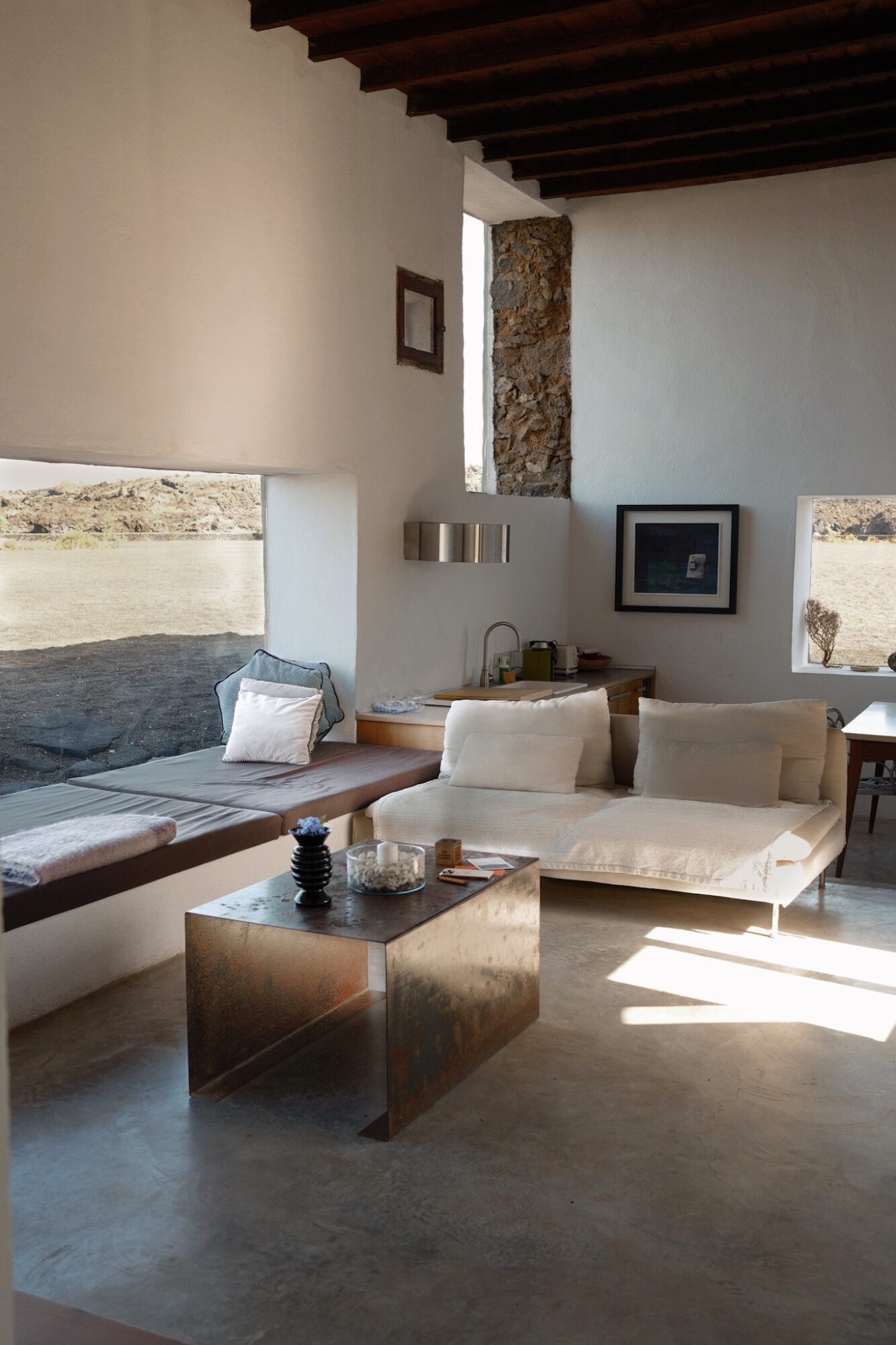 Living room with white sofas, a wooden ceiling, and windows letting in sunlight, with a stone wall visible outside.