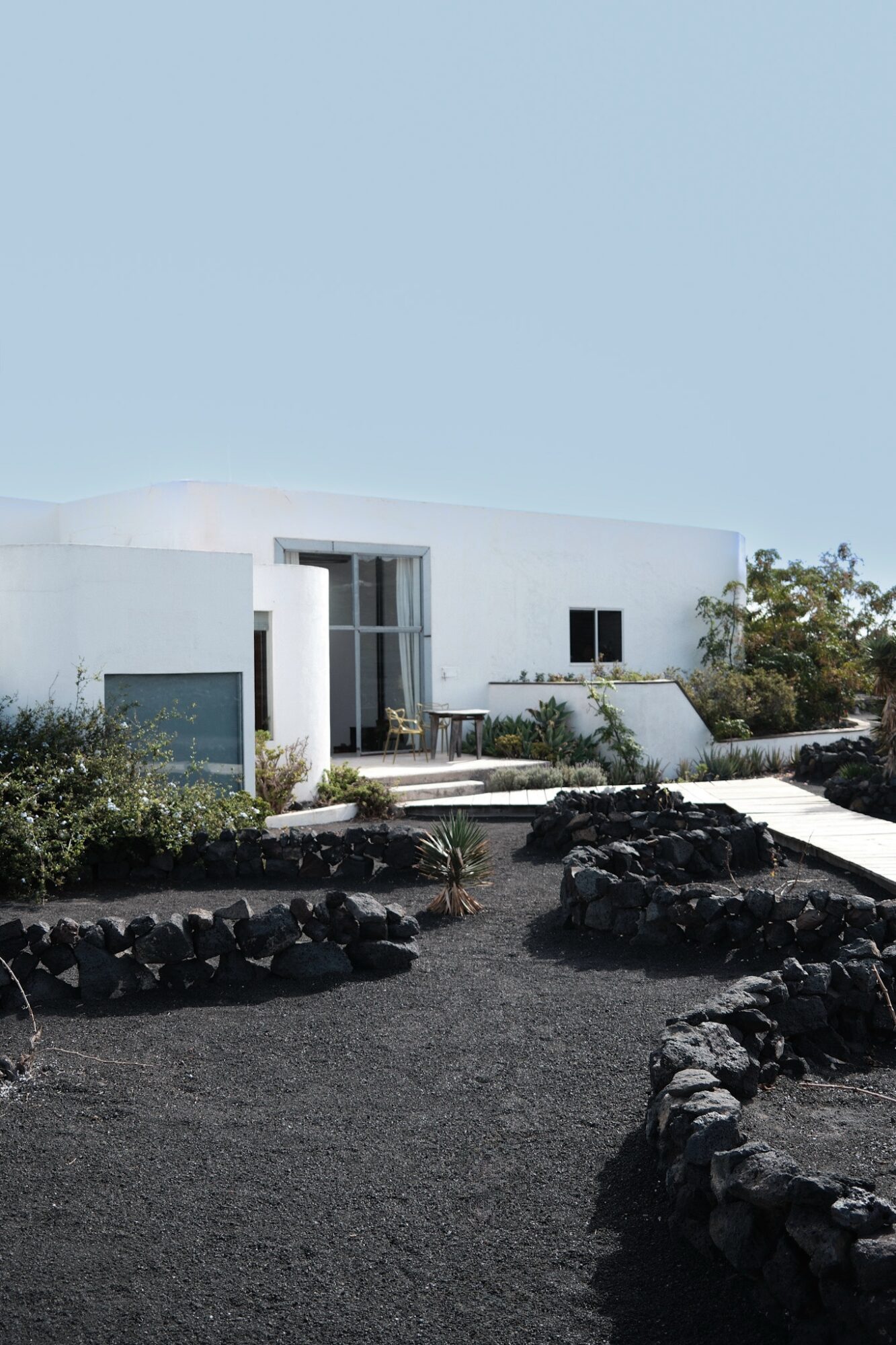 White building with a patio, surrounded by plants and black volcanic rocks, under a clear blue sky.