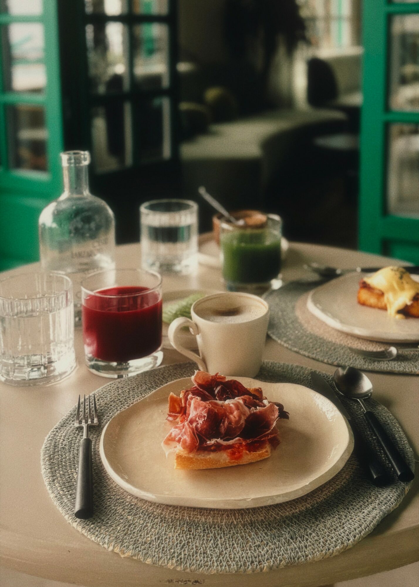 Table with a plate of toast topped with ham, a cup of coffee, a glass of red juice, and various drinks, set near a window.