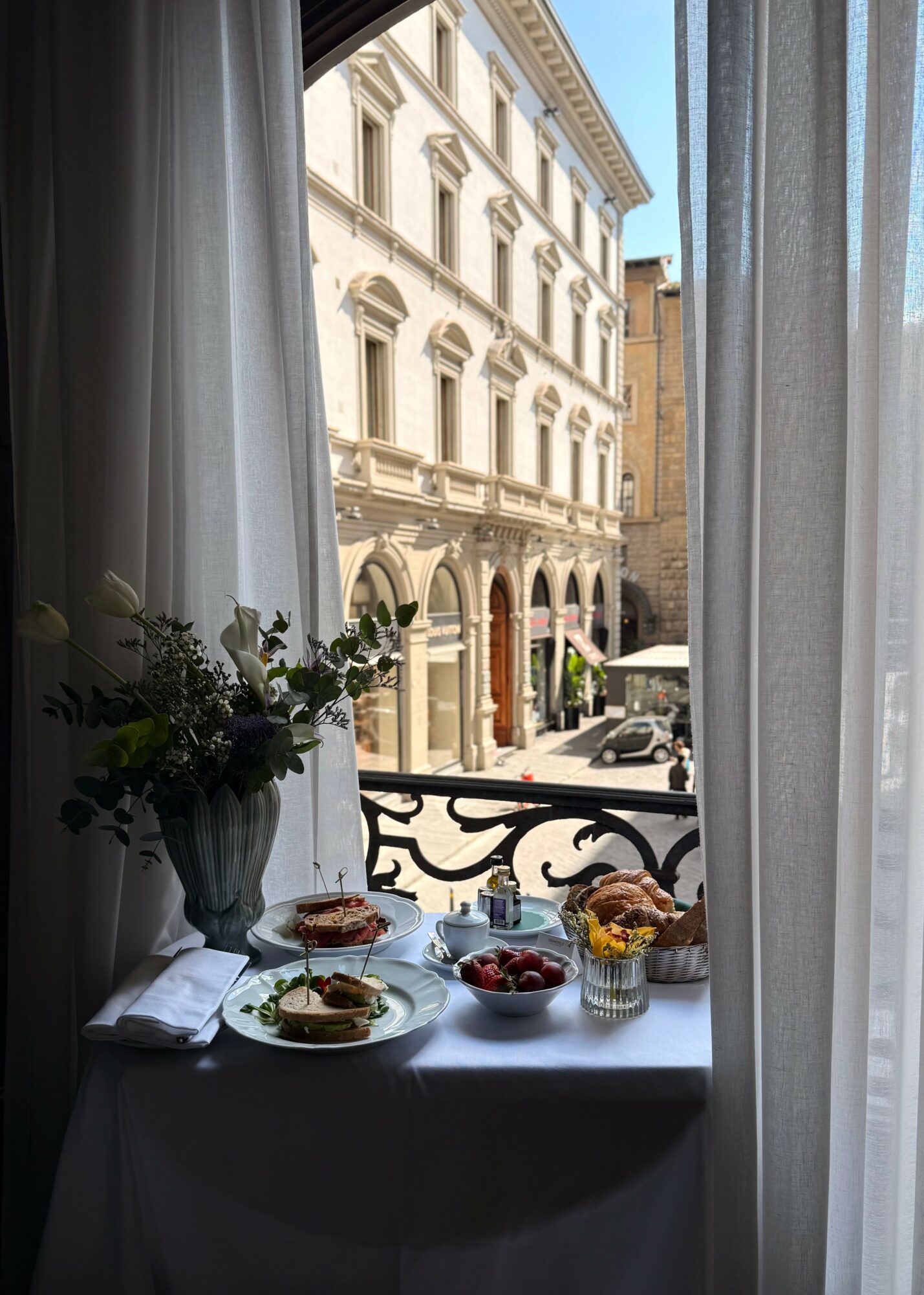 View through window with curtains showing a street scene with historic buildings and outdoor seating.
