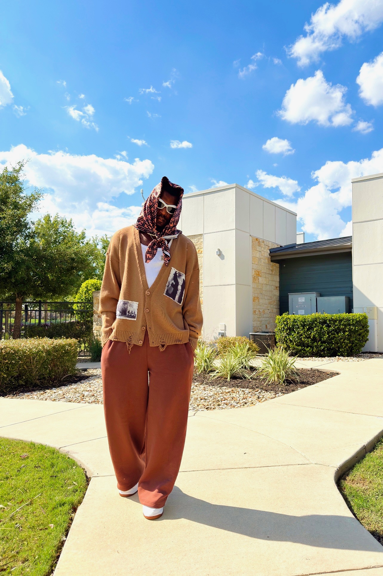 Person walking outdoors on a sunny day with blue sky and clouds, wearing a brown jacket, loose pants, and a patterned headscarf.