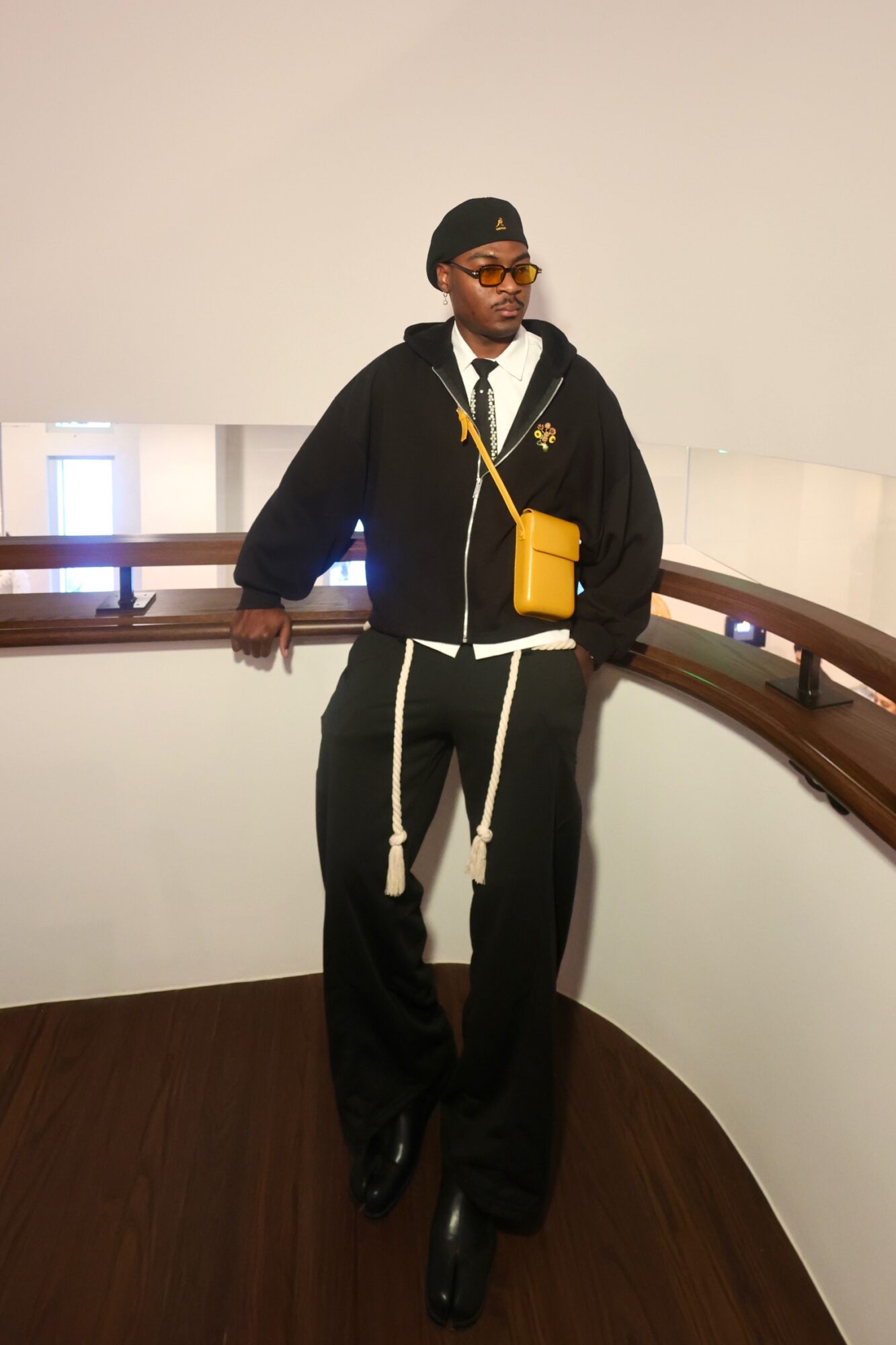 Person standing on a curved wooden floor, wearing a black jacket, black pants, and sunglasses, with a yellow bag across the chest.