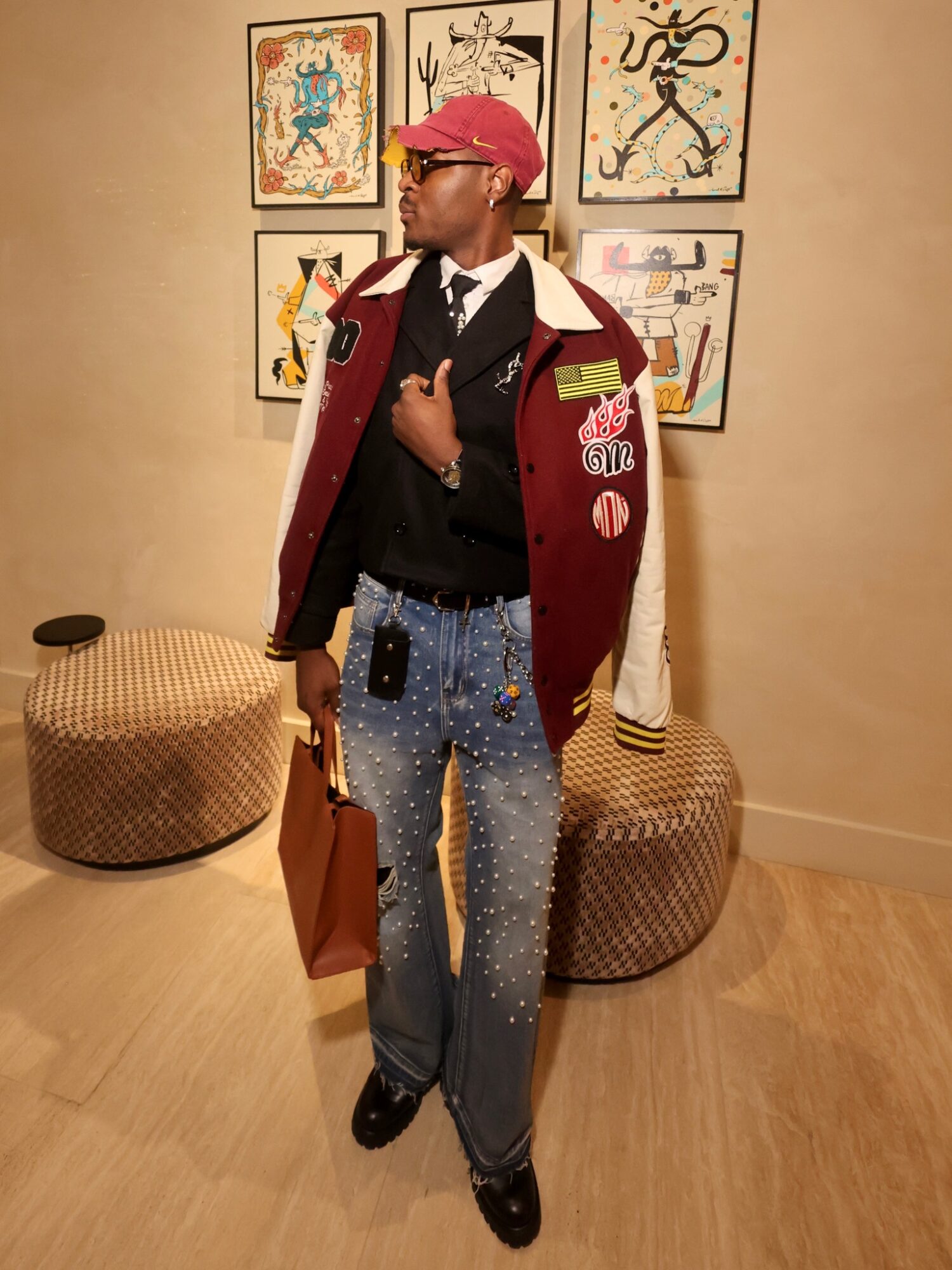 Person wearing a red hat, glasses, black jacket, and blue jeans with white paint splatters, standing in an art gallery.