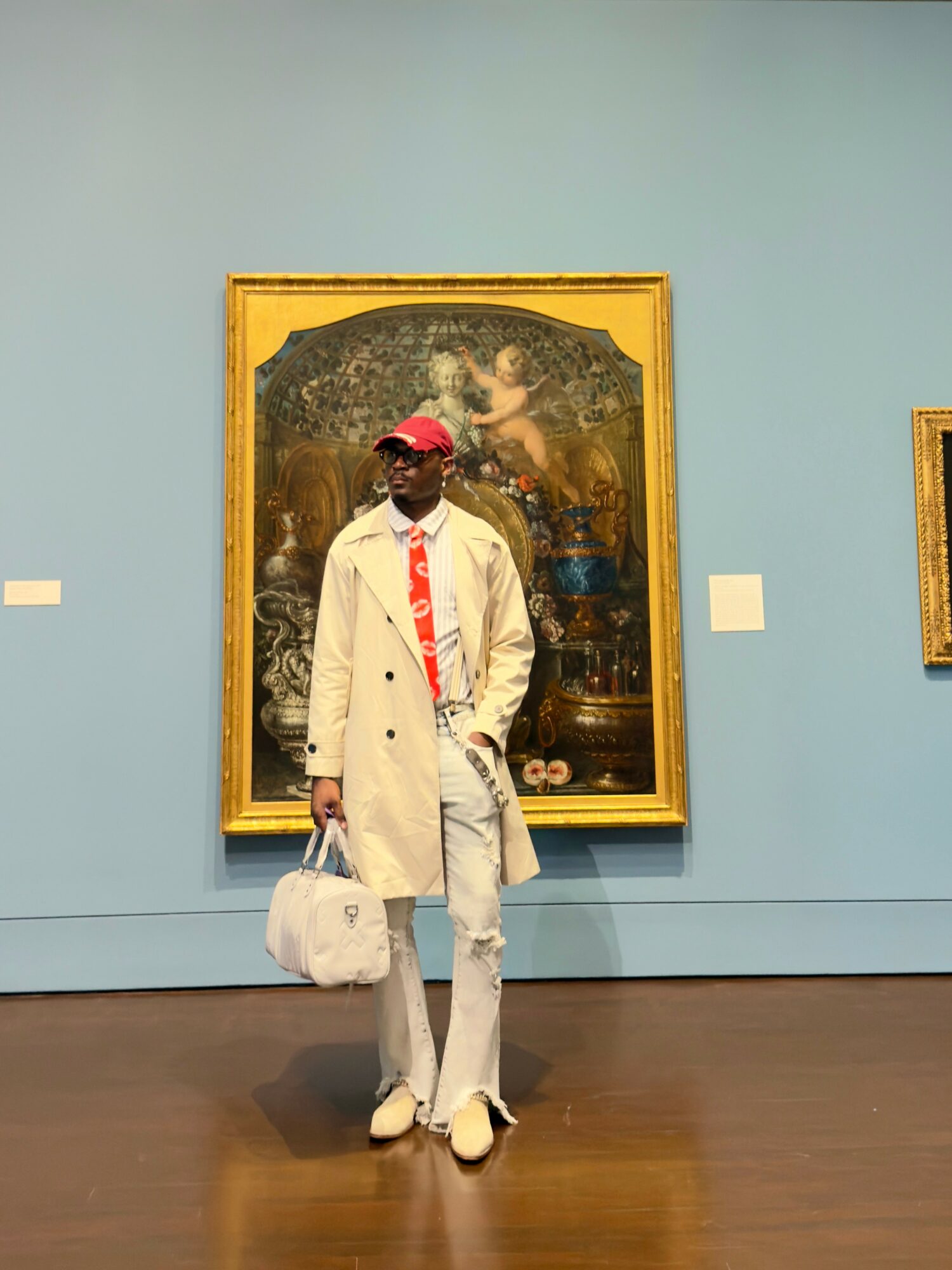Person in white coat and red cap standing in front of a large framed painting in a gallery.