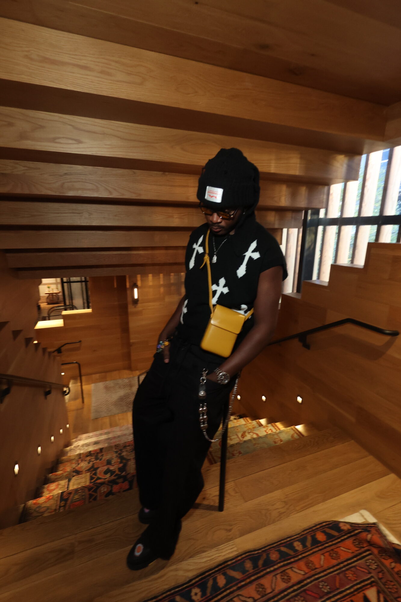 Person standing on wooden staircase, wearing black clothing, beanie, and yellow bag, in a wooden interior space.