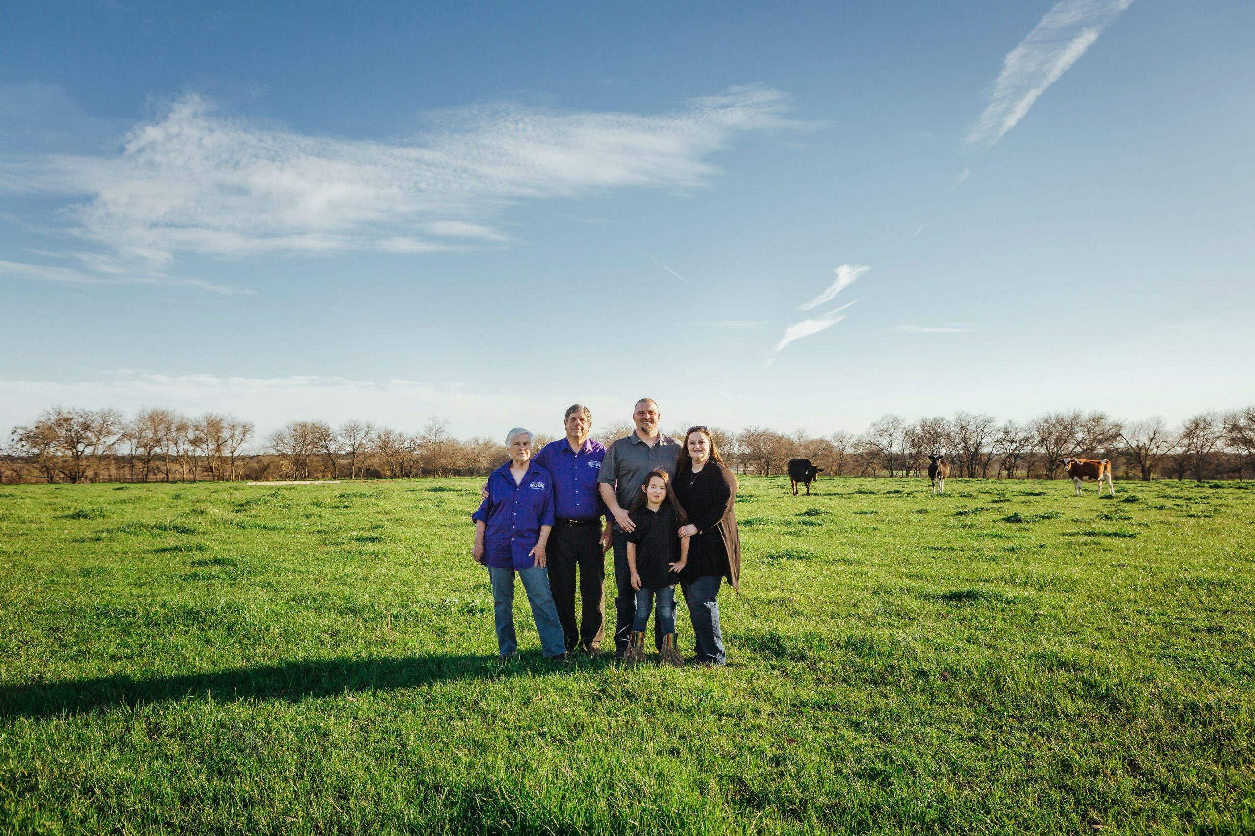 Group of five people standing in a green field with cows and trees in the background under a blue sky.