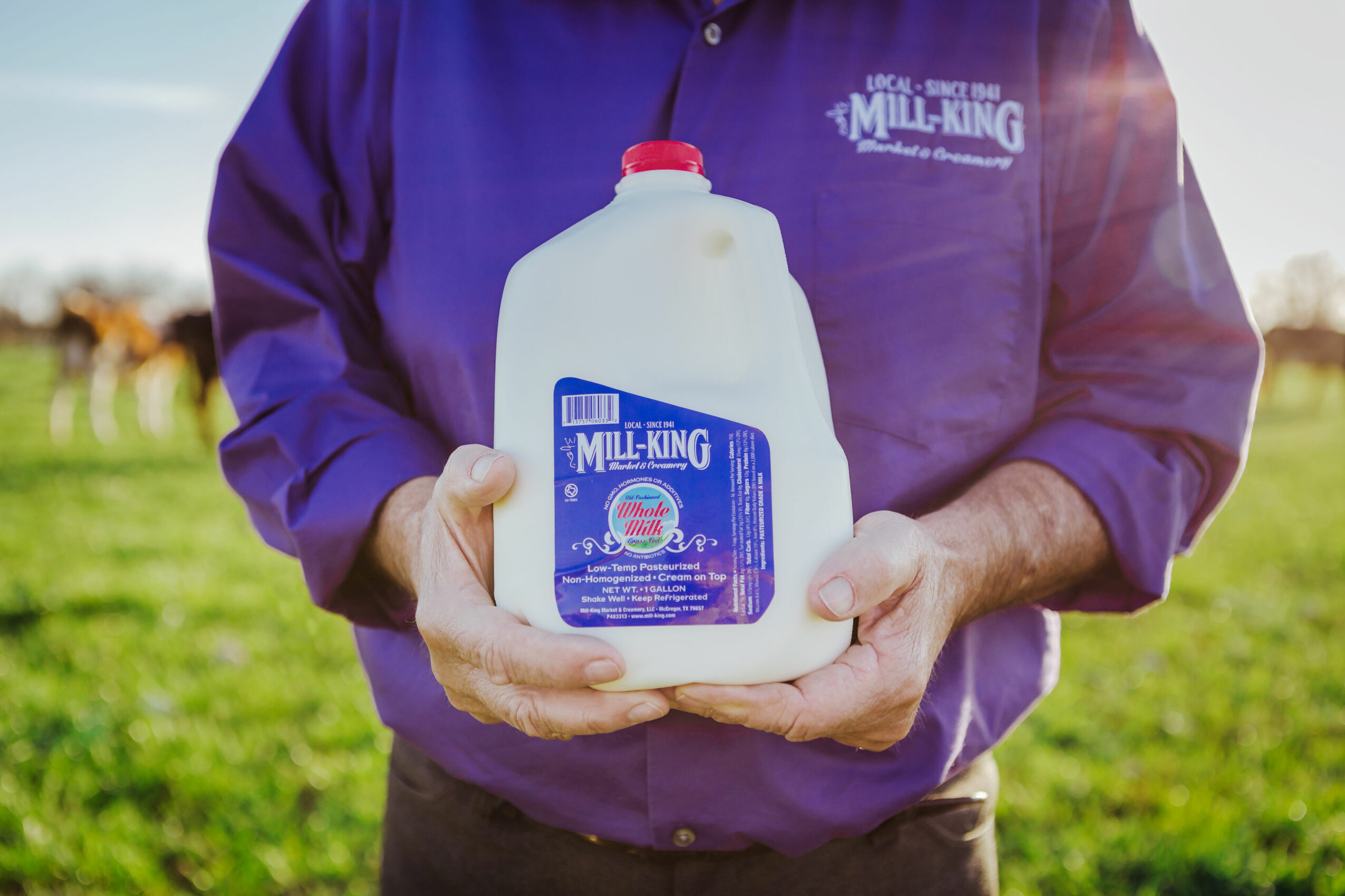 Person holding a large milk jug outdoors in a grassy field, wearing a purple shirt with a logo, sunlight in background.