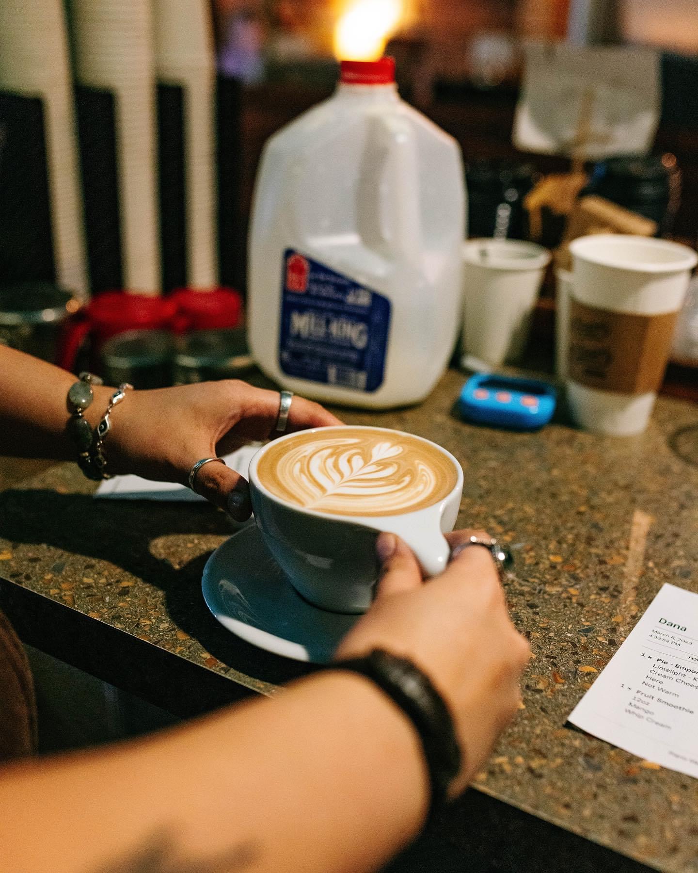 Person holding a cup of coffee with latte art, on a table with milk, cups, and a receipt.