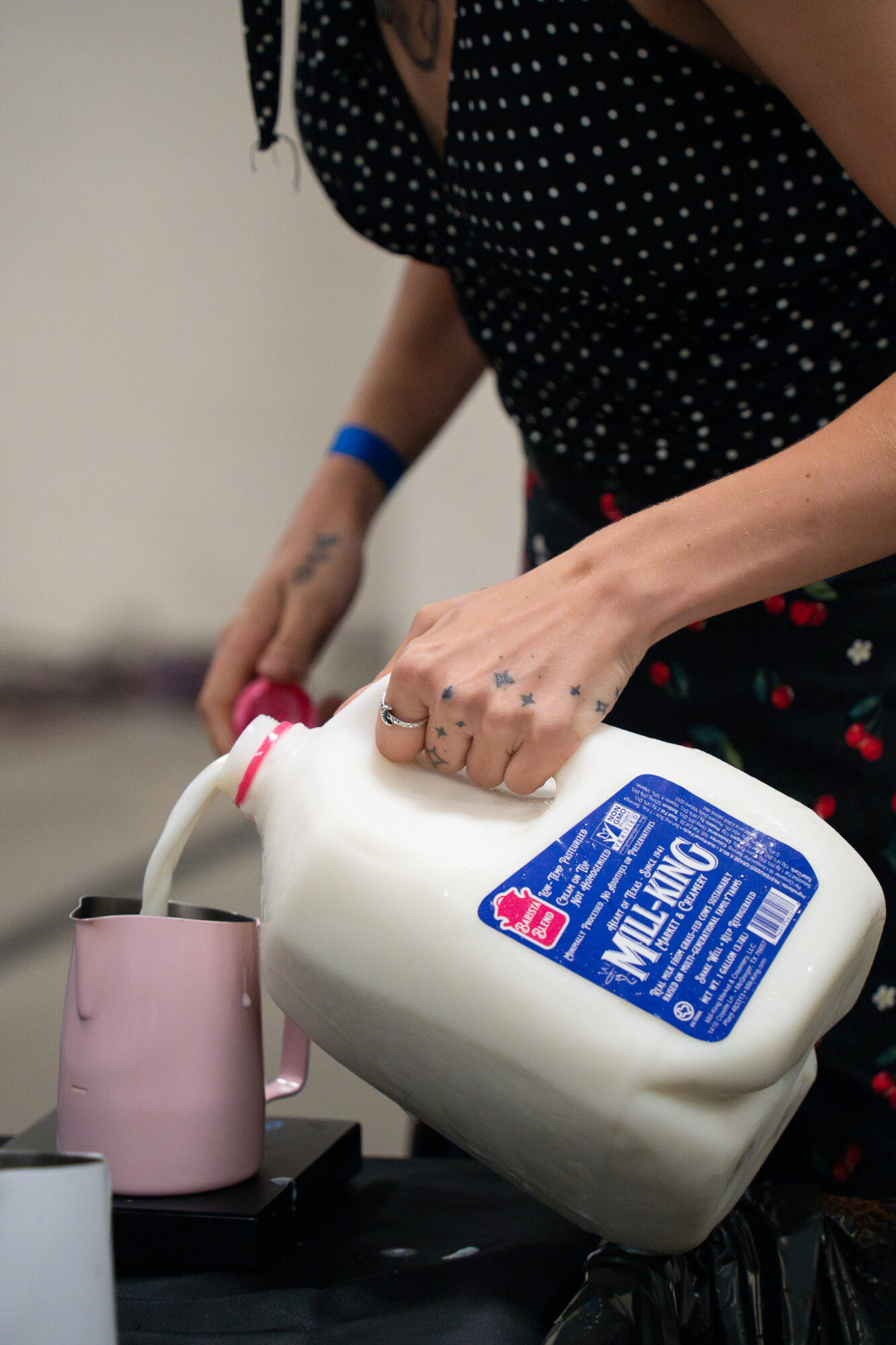 Person pouring milk from a large container into a pink cup, wearing a black polka dot shirt and a blue wristband.