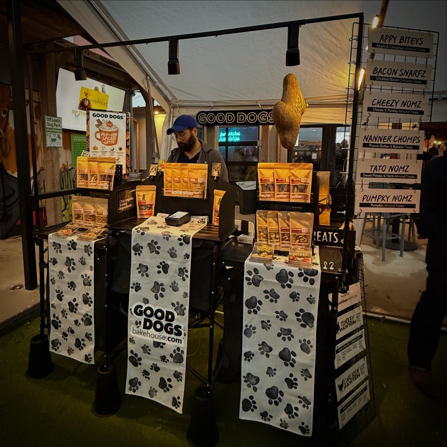 Dog treat stand with signs and a person behind the counter, inside a tented area at an event, with menu signs on the right.