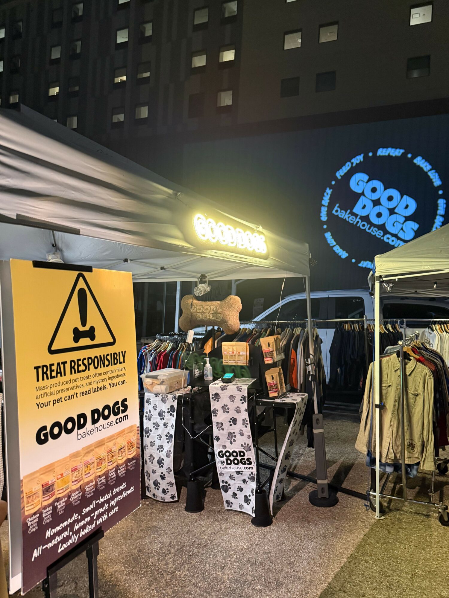 Street vendor stall with a sign, products, and a person, illuminated at night, with a building and a logo in the background.
